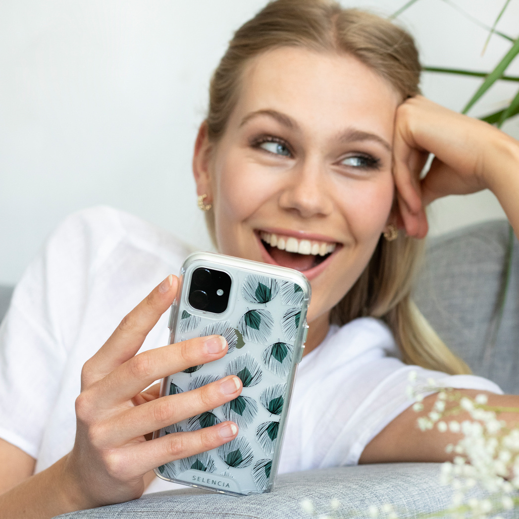 Femme blonde tenant un téléphone avec un étui à fleurs vertes. Souriante. Fond blanc.