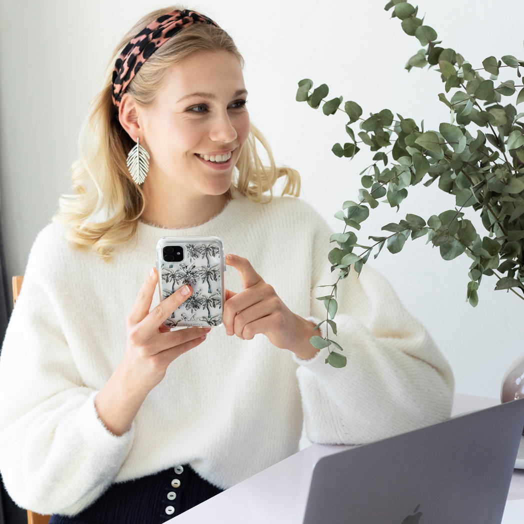 Une femme blonde tenant un téléphone avec une coque devant un ordinateur portable et une plante.