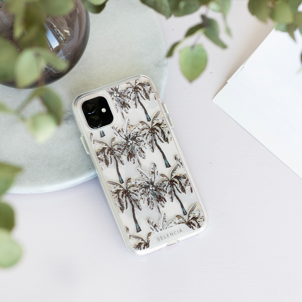 Téléphone avec étui à palmiers sur marbre, à côté d'un papier blanc et de feuilles vertes.