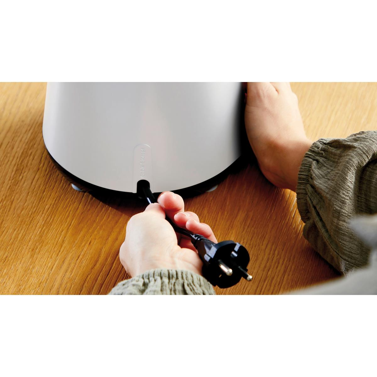 Dłonie podłączające czarny przewód zasilający do białego urządzenia, na drewnianej powierzchni.