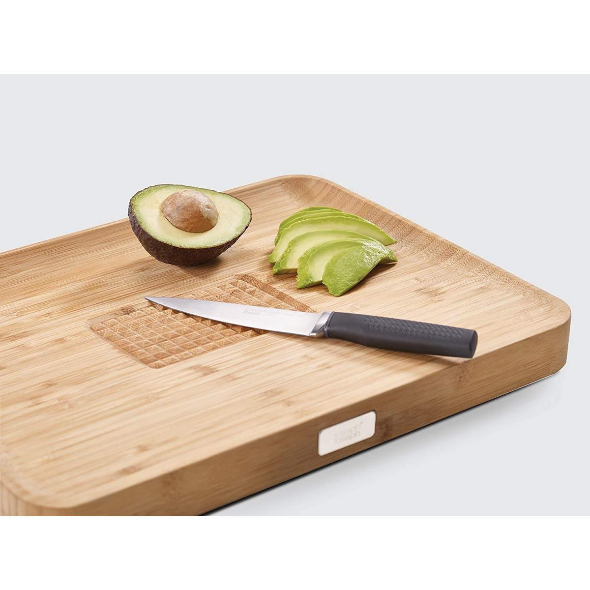 Holzschneidebrett mit Avocadohälfte und Scheiben sowie einem kleinen Messer. Avocado und Messer befinden sich auf einem gemusterten Bereich.
