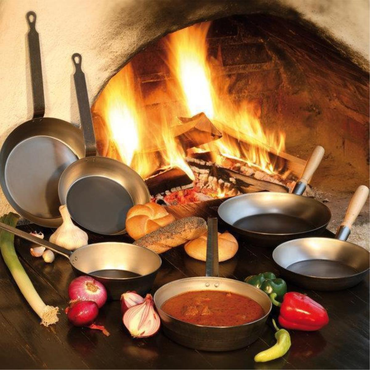 Mehrere Pfannen mit Essen und Zutaten vor einem Kamin mit brennendem Holz.
