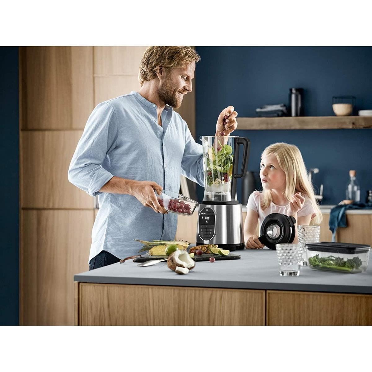 Hombre y niña usando una licuadora, ingredientes y un vaso de batido en la encimera.