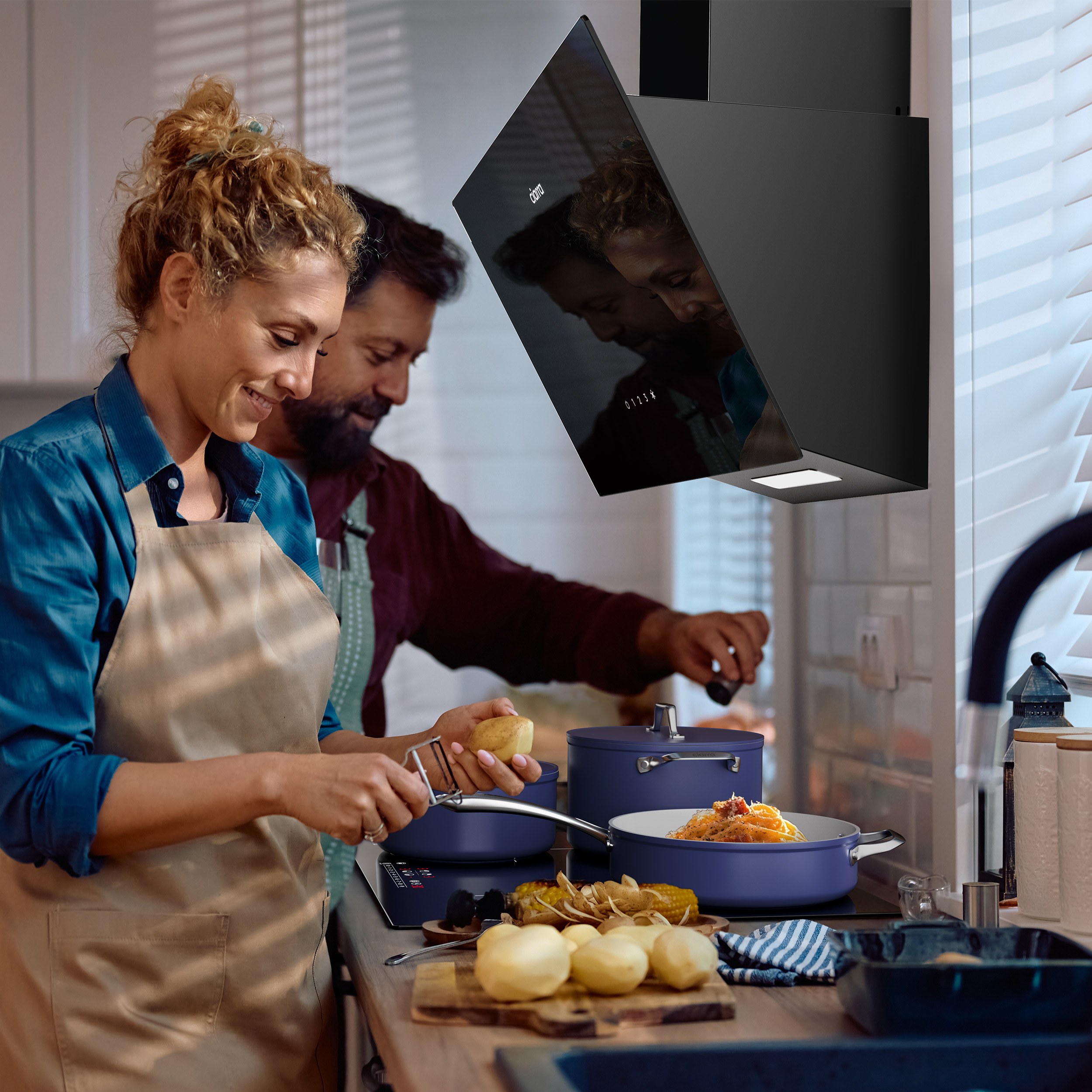 Koppel in een keuken. Vrouw schilt aardappelen, man kookt. Een zwarte afzuigkap boven het fornuis.