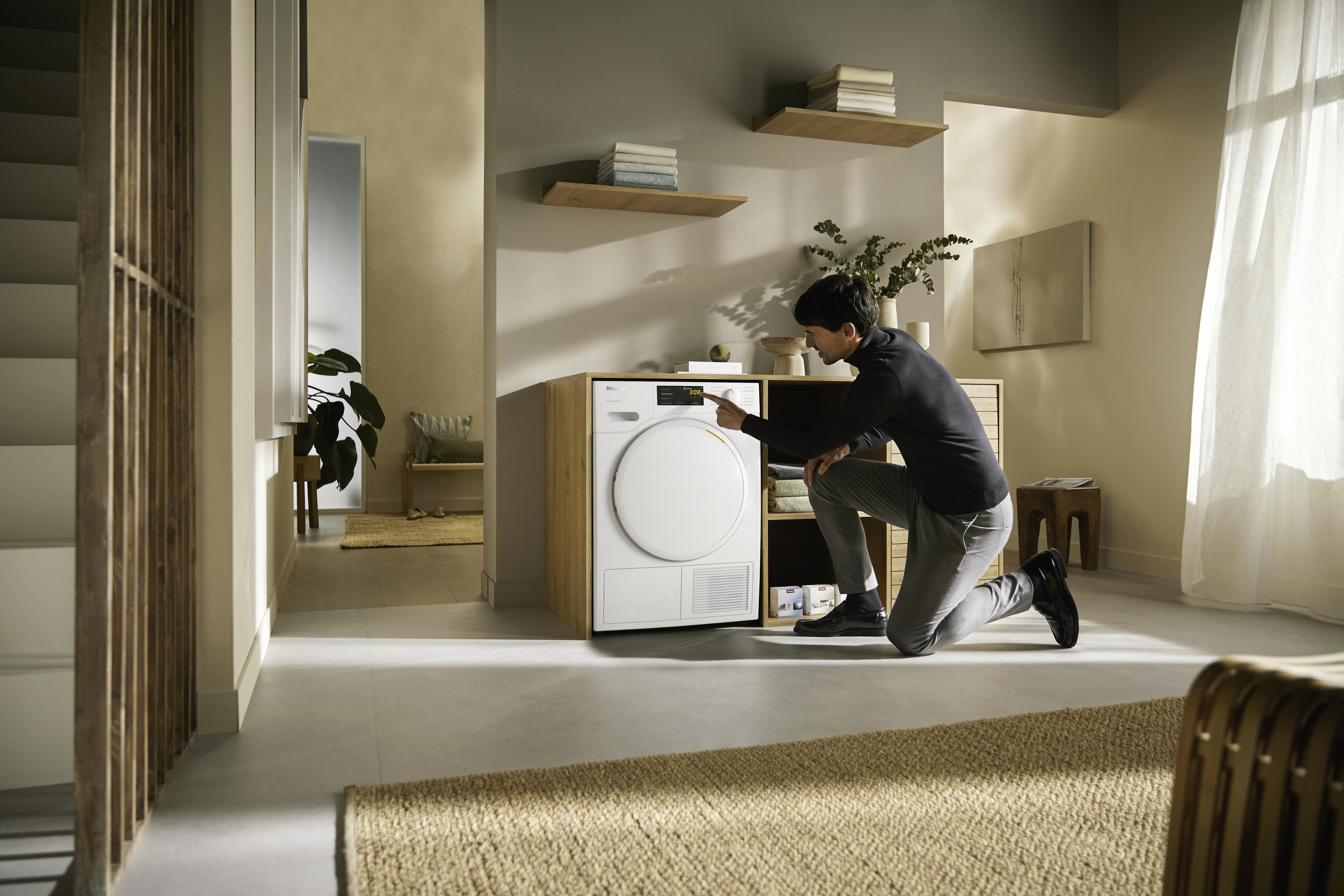 Homme à genoux dans une pièce moderne, interagissant avec un lave-linge blanc. Le fond est un mur et des étagères.