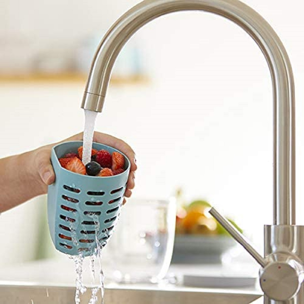 Eine Hand hält ein blaues Sieb unter einem Wasserhahn. Erdbeeren und Blaubeeren sind im Sieb. Wasser läuft.