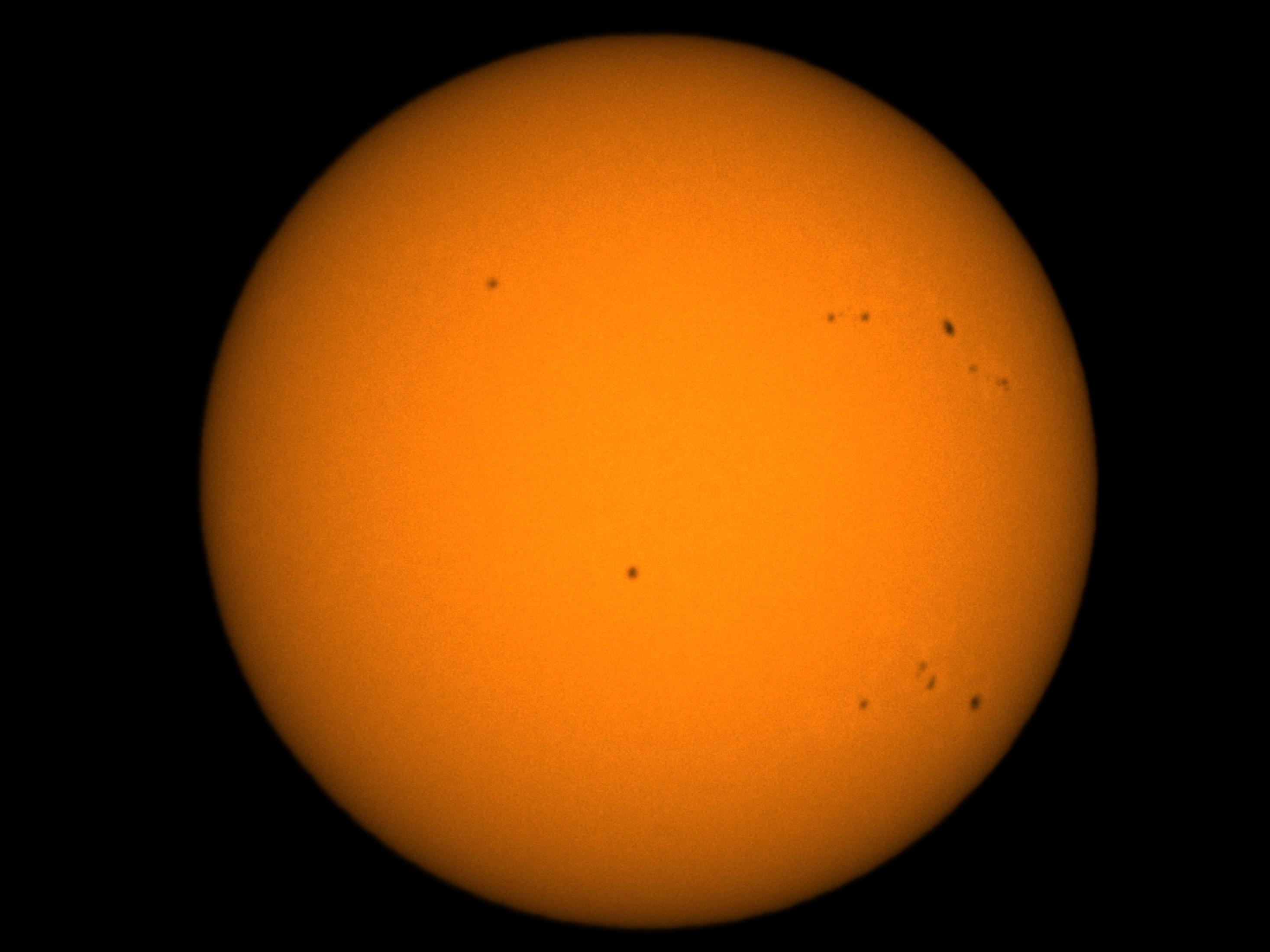 Die Sonne, eine orangefarbene Kugel mit dunklen Flecken auf schwarzem Hintergrund. Die Sonne scheint im Fokus zu stehen.