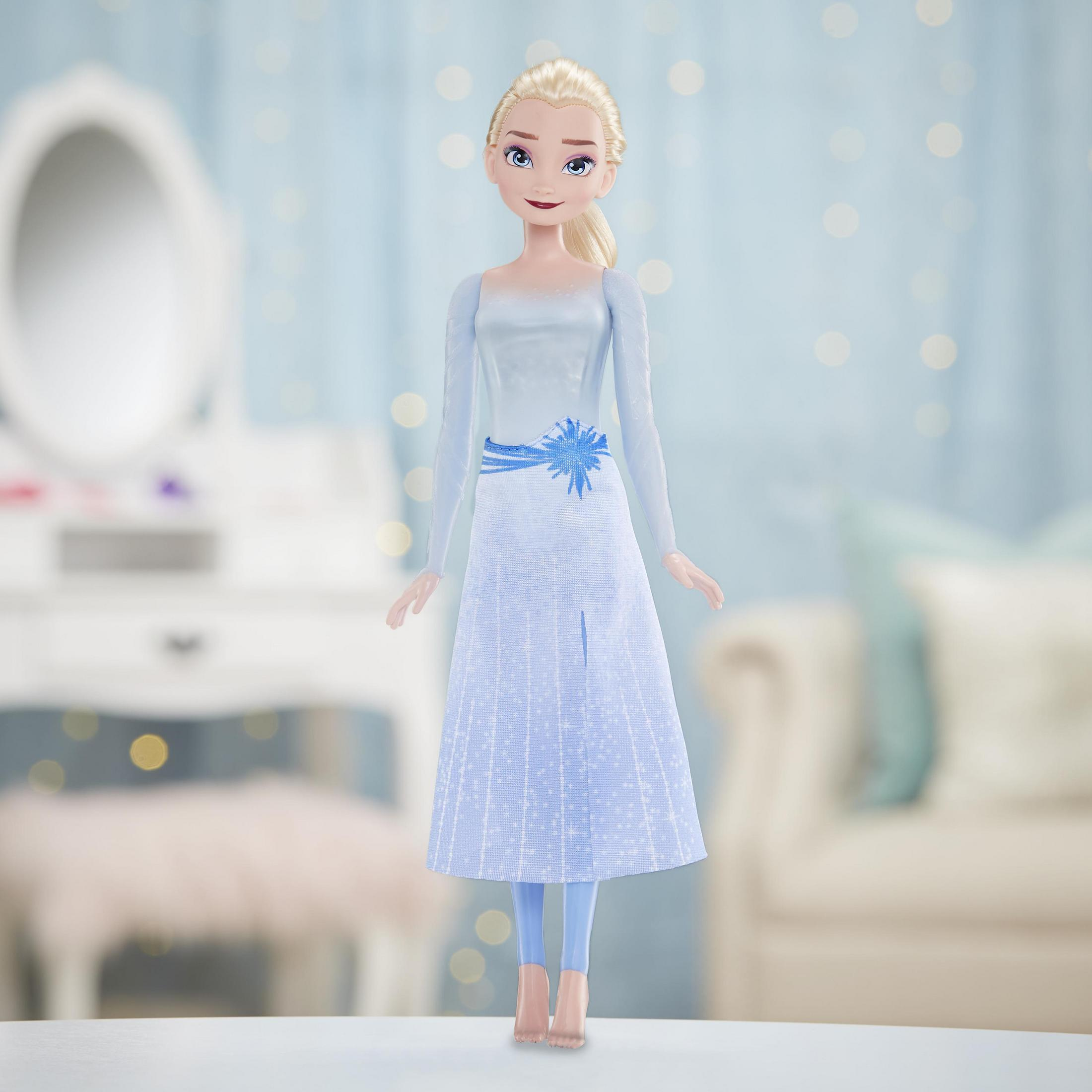 Elsa-Puppe in blauem Kleid. Blondes Haar, steht in einem Raum mit Schminktisch und Sofa.