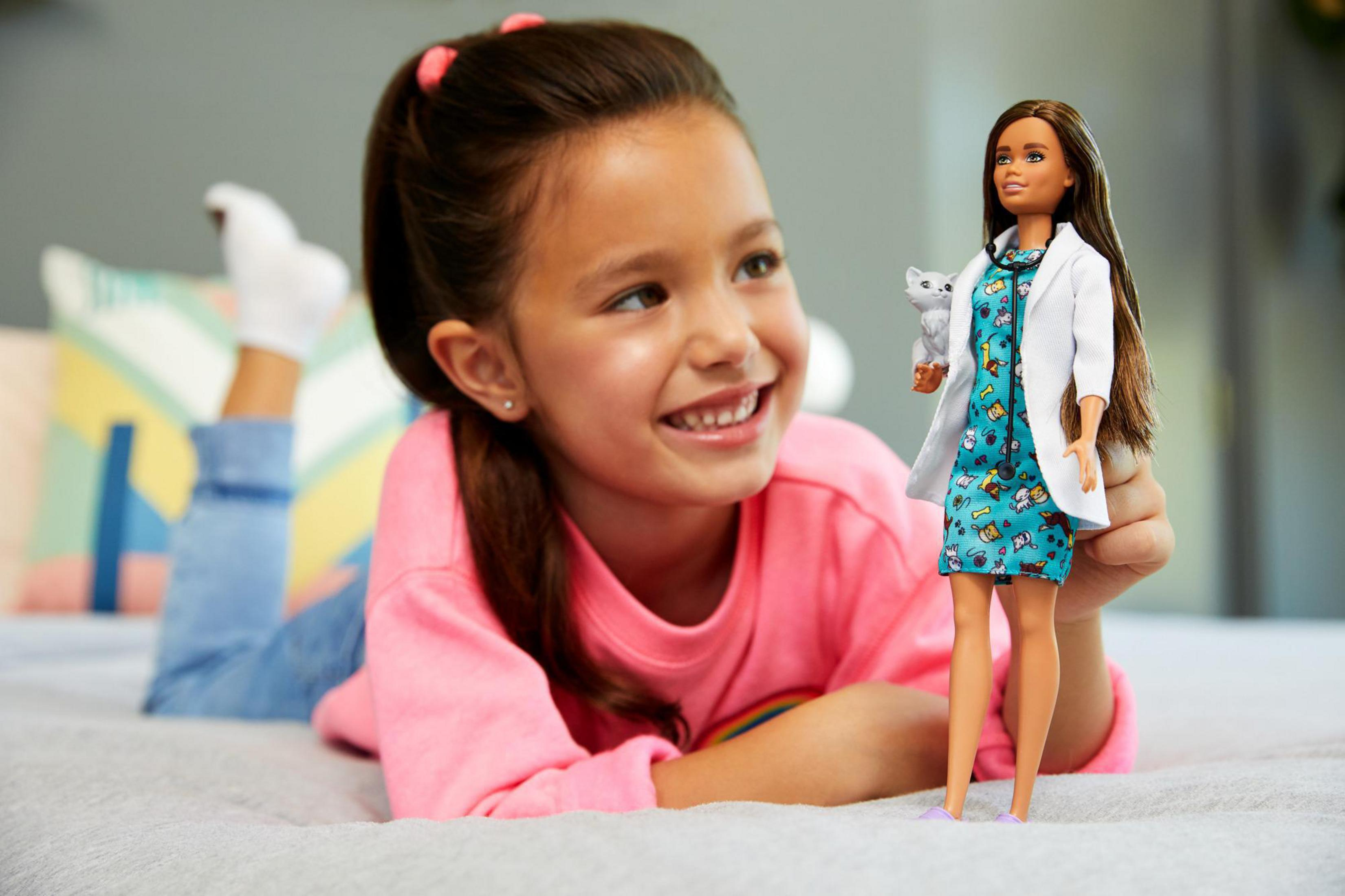 Mädchen hält eine Barbie-Puppe als Tierärztin. Lächelt, liegt auf einem Bett.