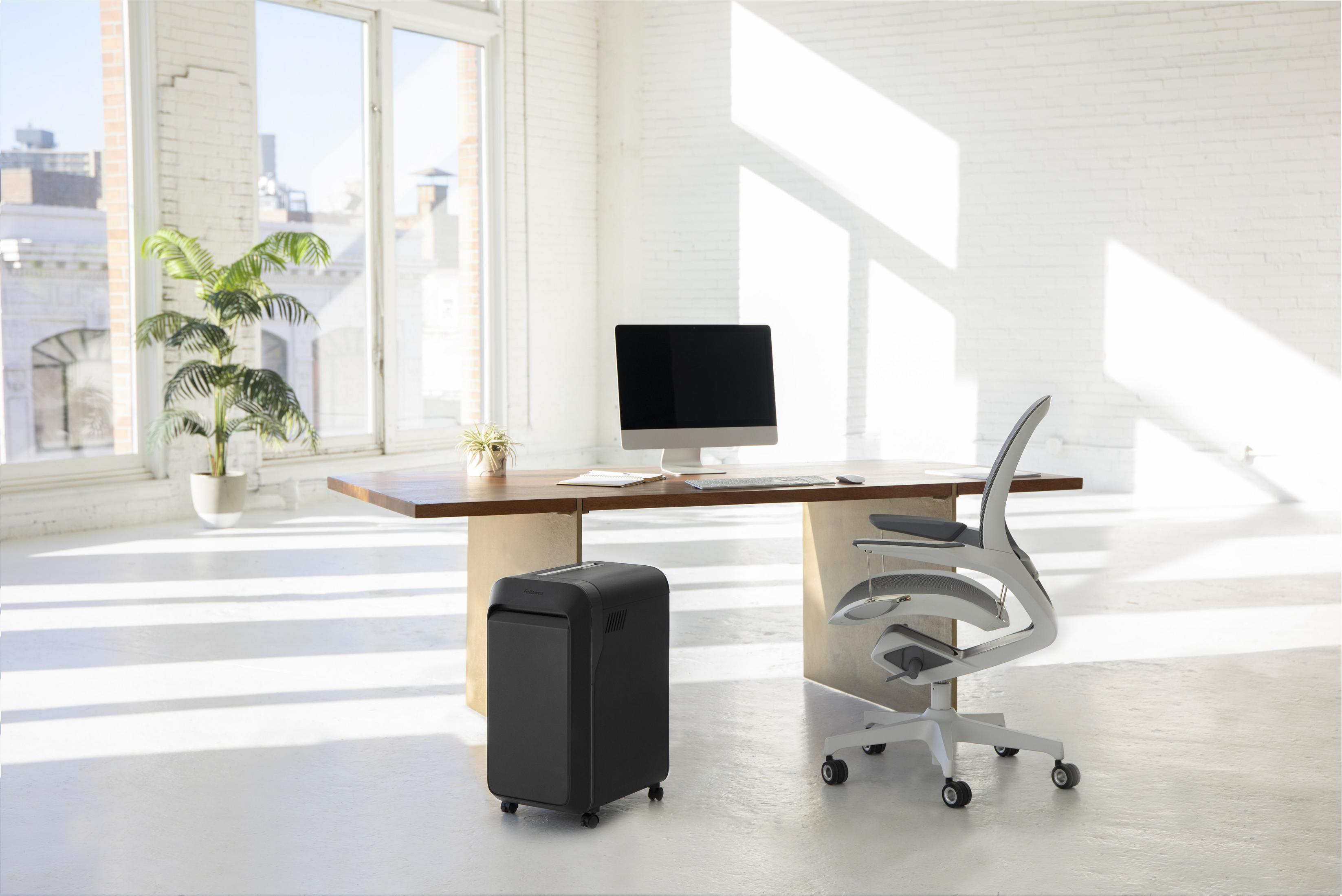 Modern office with a desk, computer, shredder and a white ergonomic chair near a window.