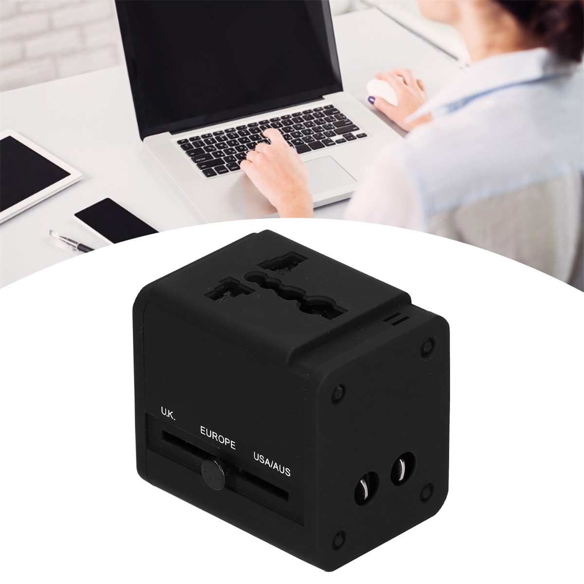 Ein schwarzer Universal-Reiseadapter. Im Hintergrund arbeitet eine Person an einem Laptop.