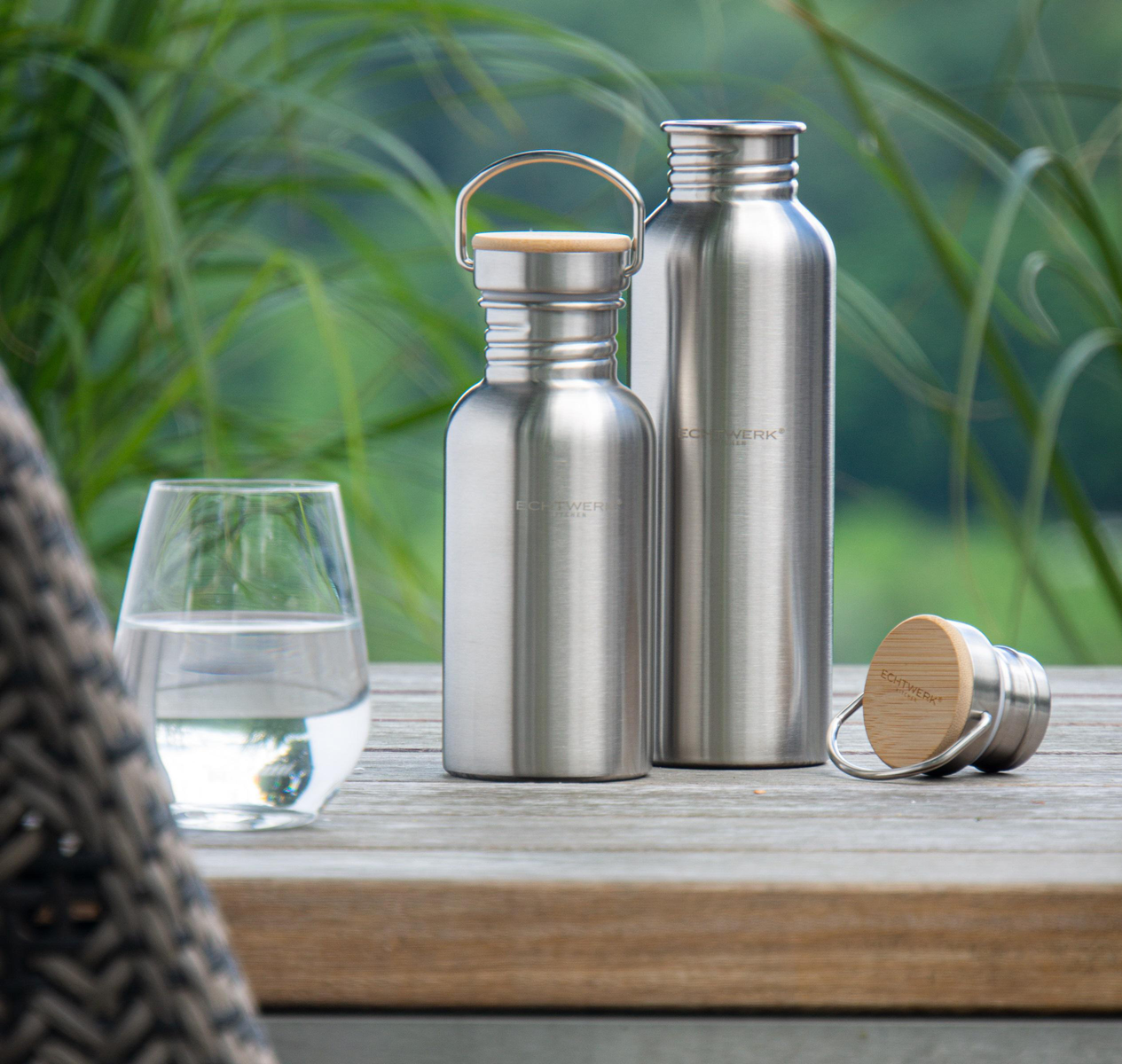 Zwei Edelstahl-Wasserflaschen mit Holzkappen und ein Glas Wasser auf Holzoberfläche.