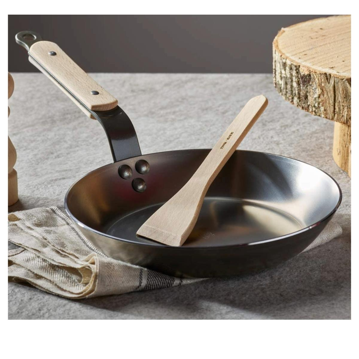 Eine Edelstahlpfanne mit einem Holzspatel. Ein Holzscheit und ein weißer Hintergrund.