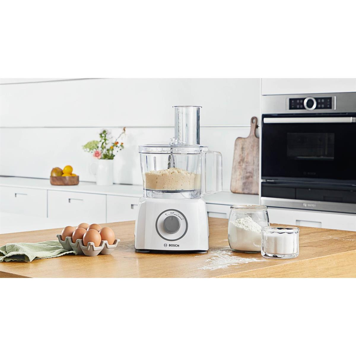 Biały robot kuchenny z ciastem, jajkami i pojemnikami na drewnianym blacie w nowoczesnej kuchni.