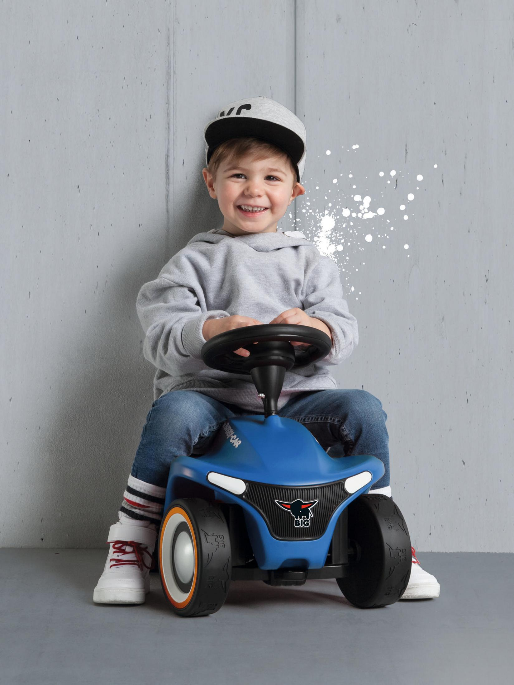 Un niño pequeño sonriendo mientras está sentado en un coche de juguete azul contra una pared gris.