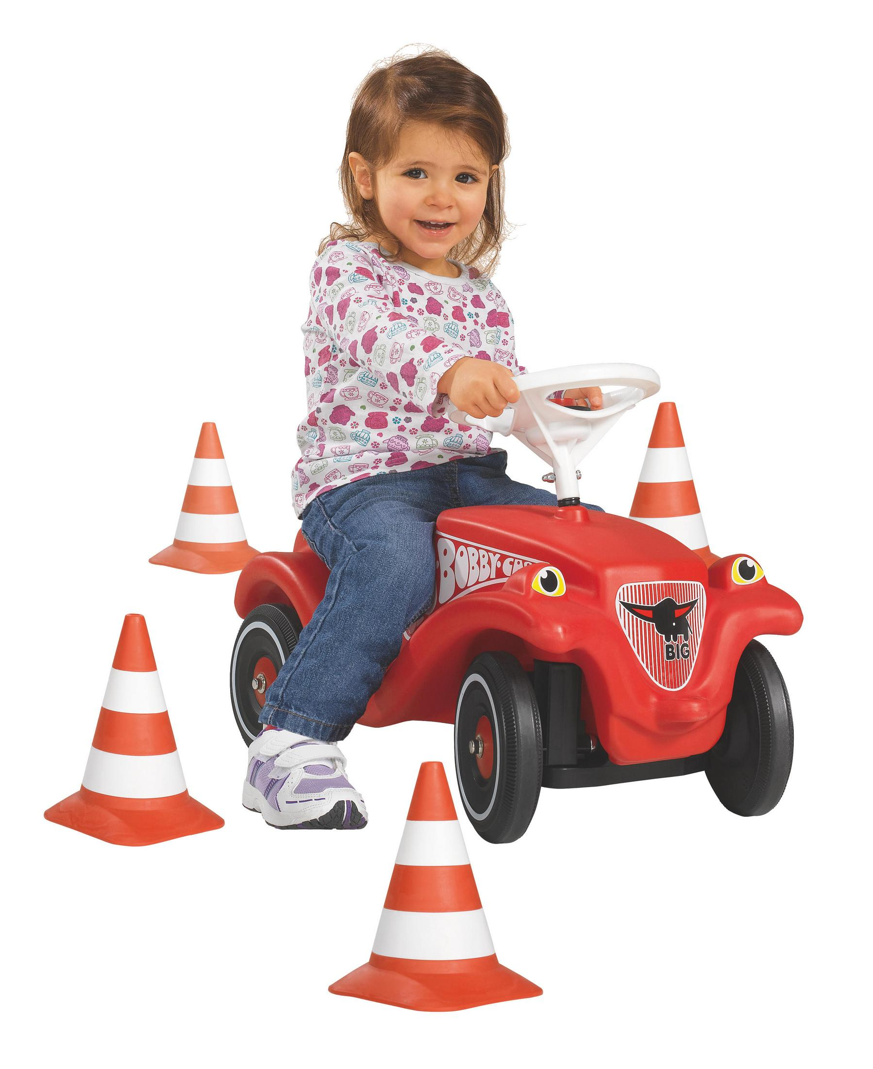 Ein junges Mädchen fährt ein rotes Spielzeugauto. Verkehrskegel umgeben das Auto. Weißer Hintergrund.