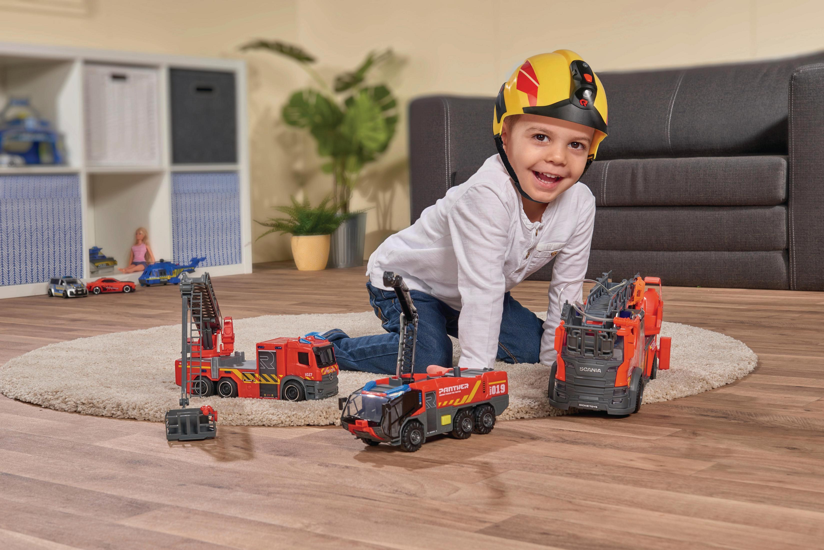 Dziecko w kasku bawi się zabawkowymi wozami strażackimi na dywanie. Zabawki są czerwone i pomarańczowe.
