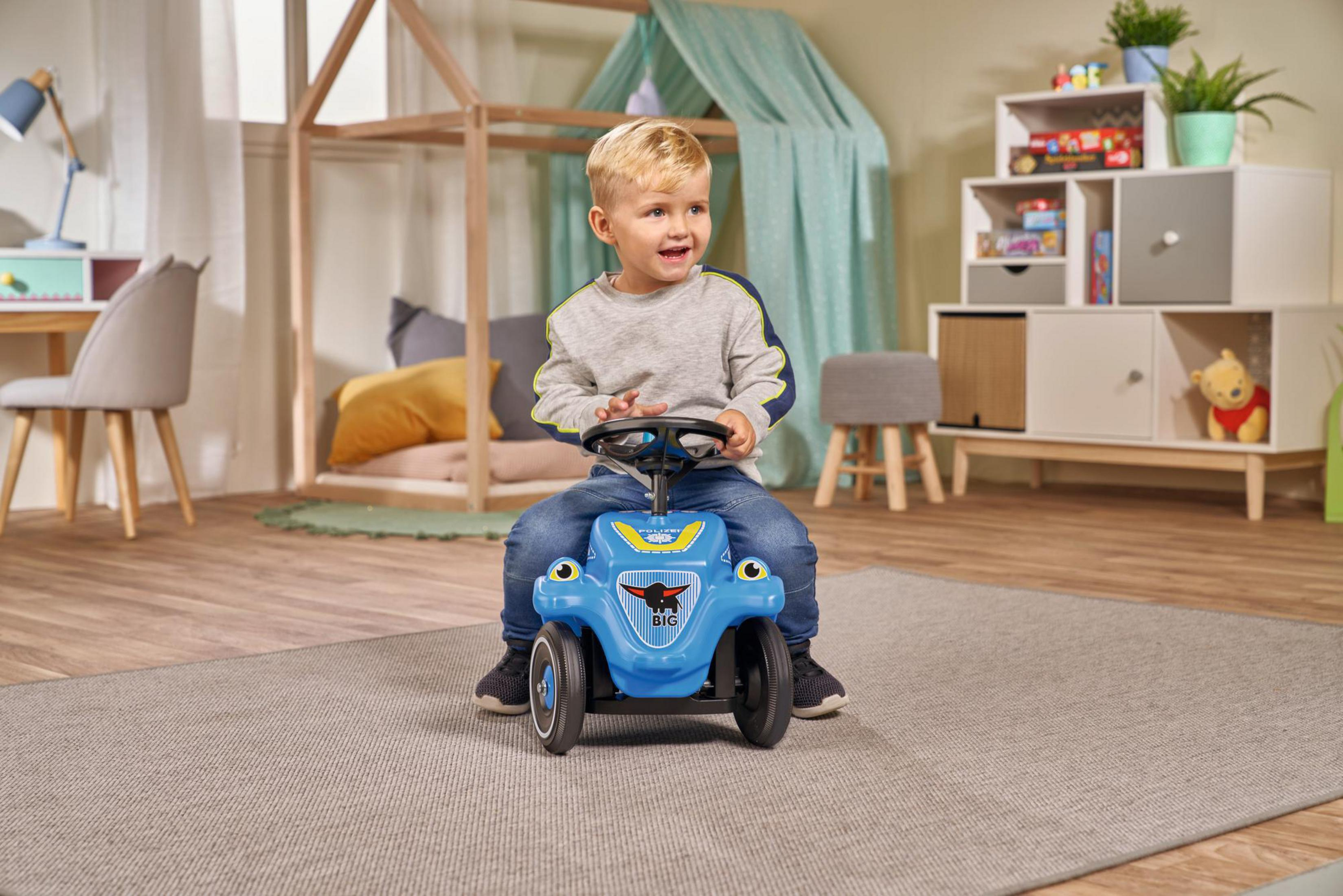 Kleiner Junge auf einem blauen Spielzeugauto, auf einem grauen Teppich in einem Raum mit Holzmöbeln.