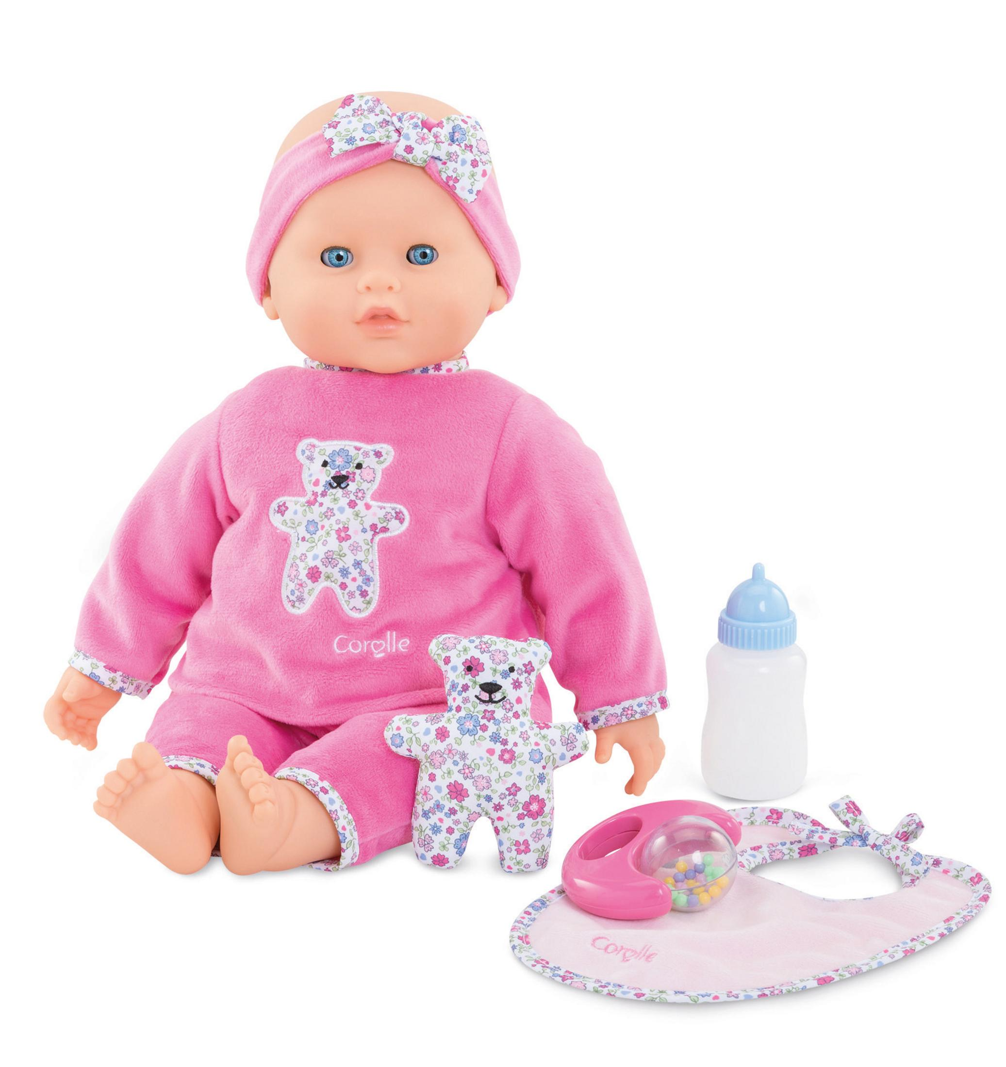 Eine Babypuppe mit rosa Outfit, einem Teddybär, einer Flasche und einem Lätzchen. Alle Artikel sind auf weißem Untergrund.