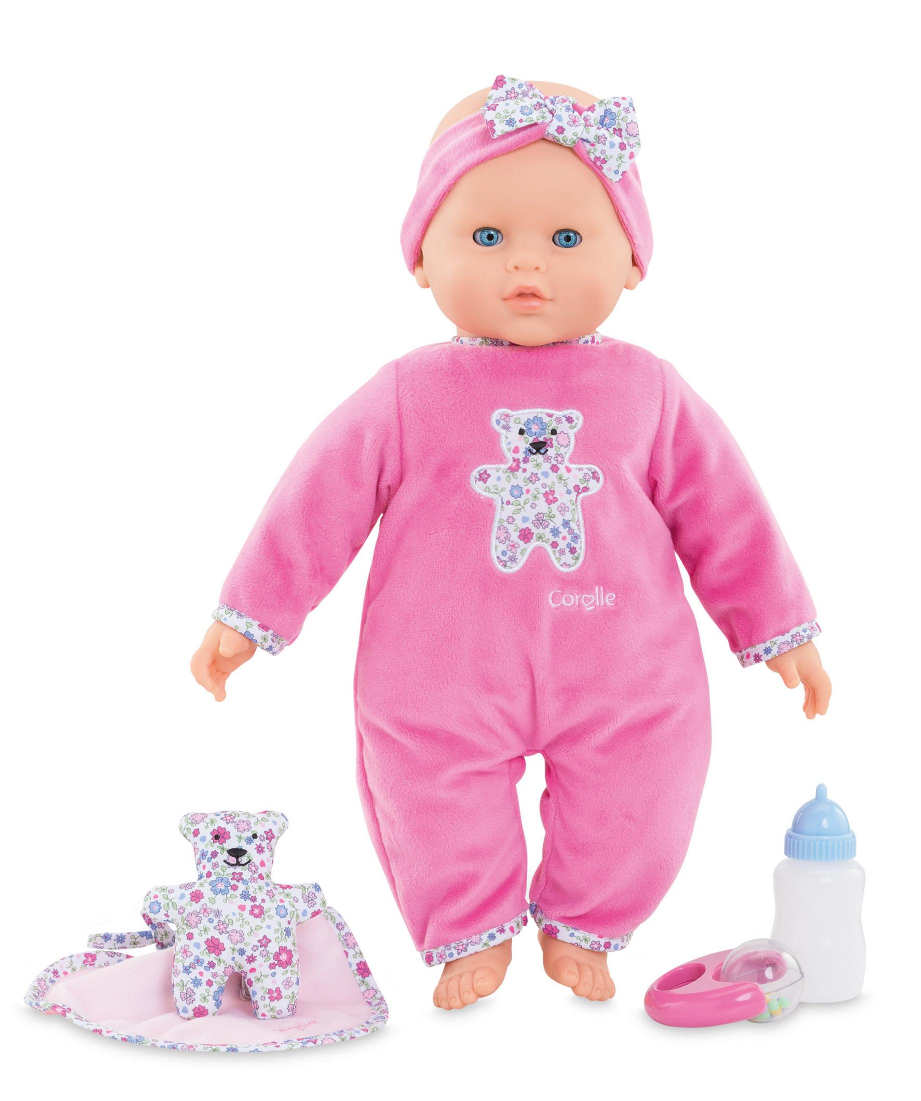 Eine Babypuppe in rosa Pyjama und Stirnband, mit einem Bärenplüsch, einer Decke und einer Flasche.