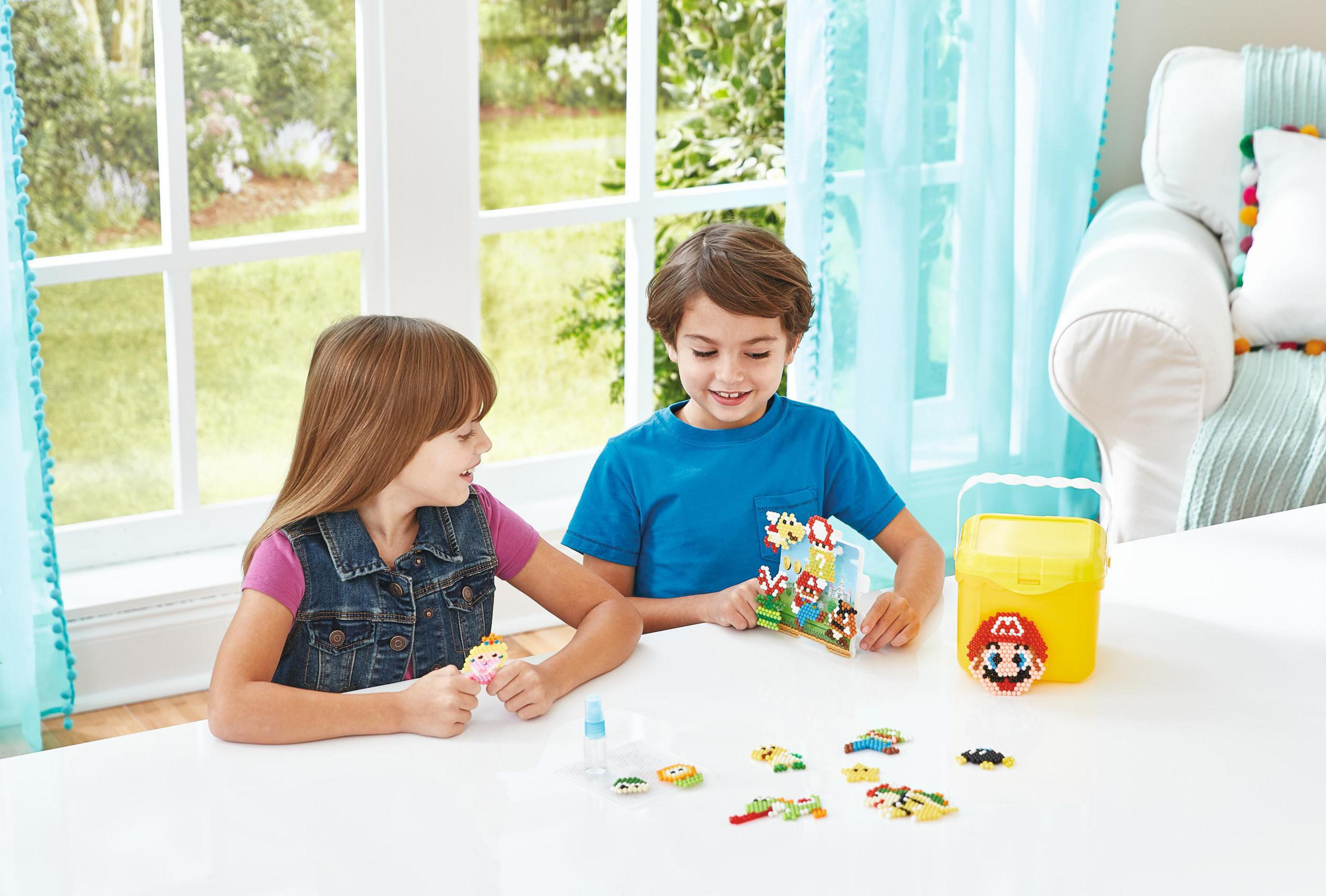 Due bambini a un tavolo con perline colorate e un'esposizione di Super Mario. Un secchio giallo e una finestra sullo sfondo.