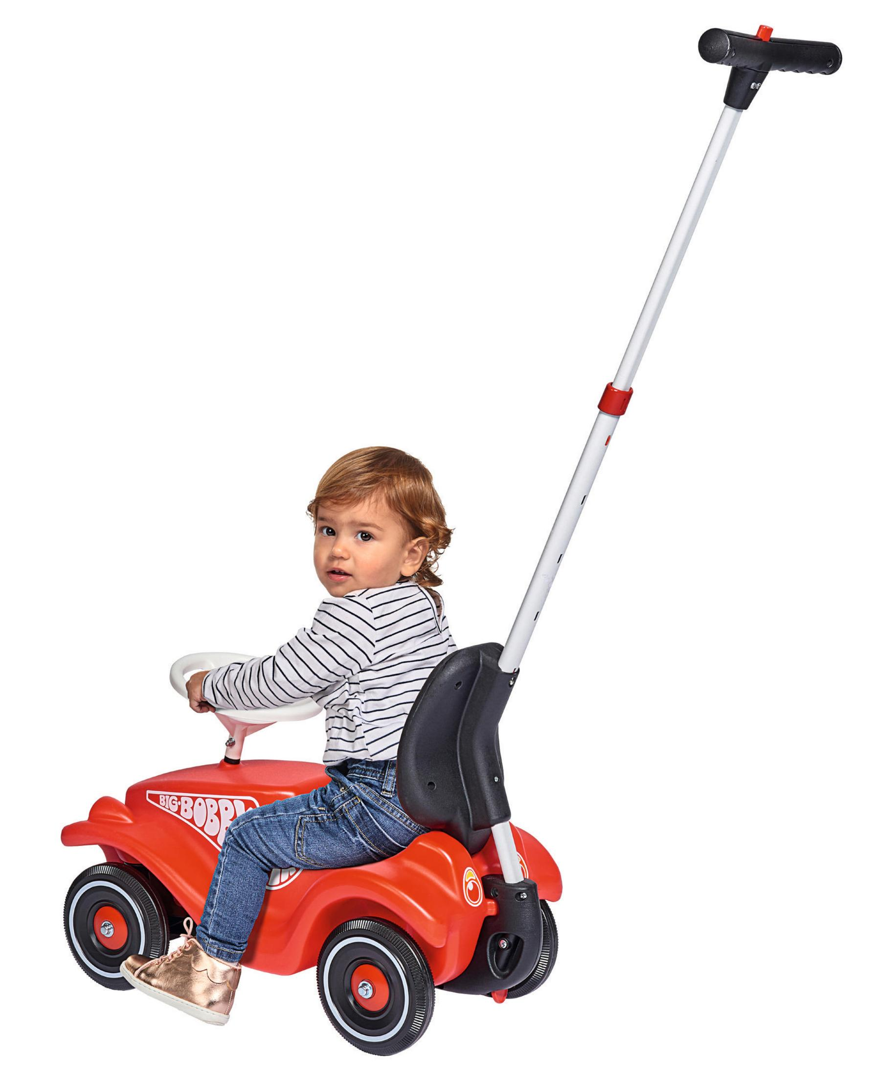 Dziecko siedzi w czerwonym samochodziku z uchwytem dla rodziców. Dziecko jest blondynem.