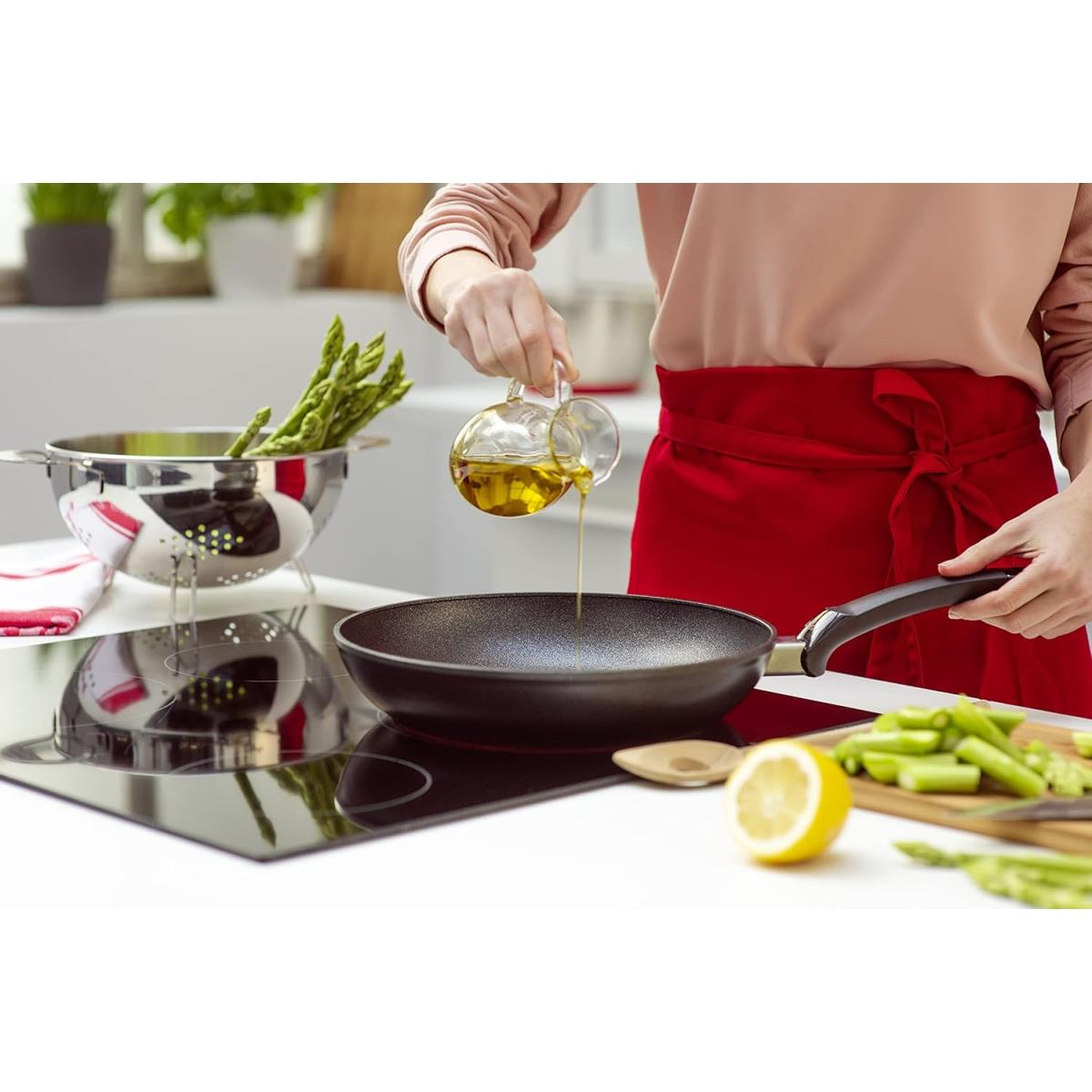 Una persona versa olio in una padella nera su un fornello, con asparagi e limone nelle vicinanze.