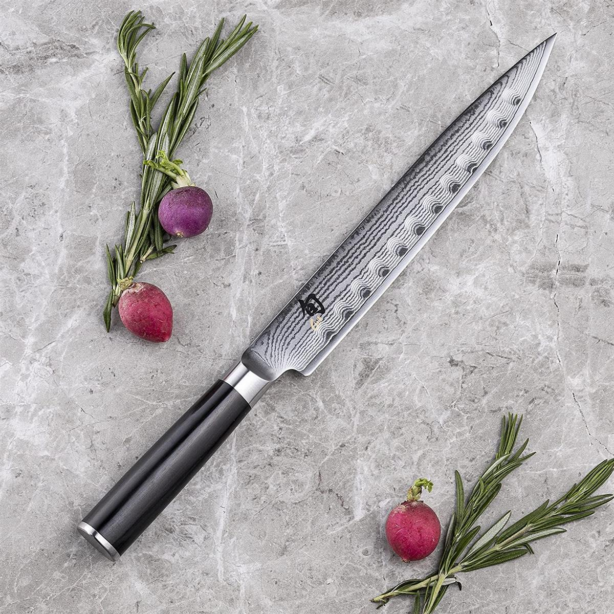 Ein langes, silbernes Messer mit dunklem Griff liegt auf Stein mit Kräutern und Radieschen.