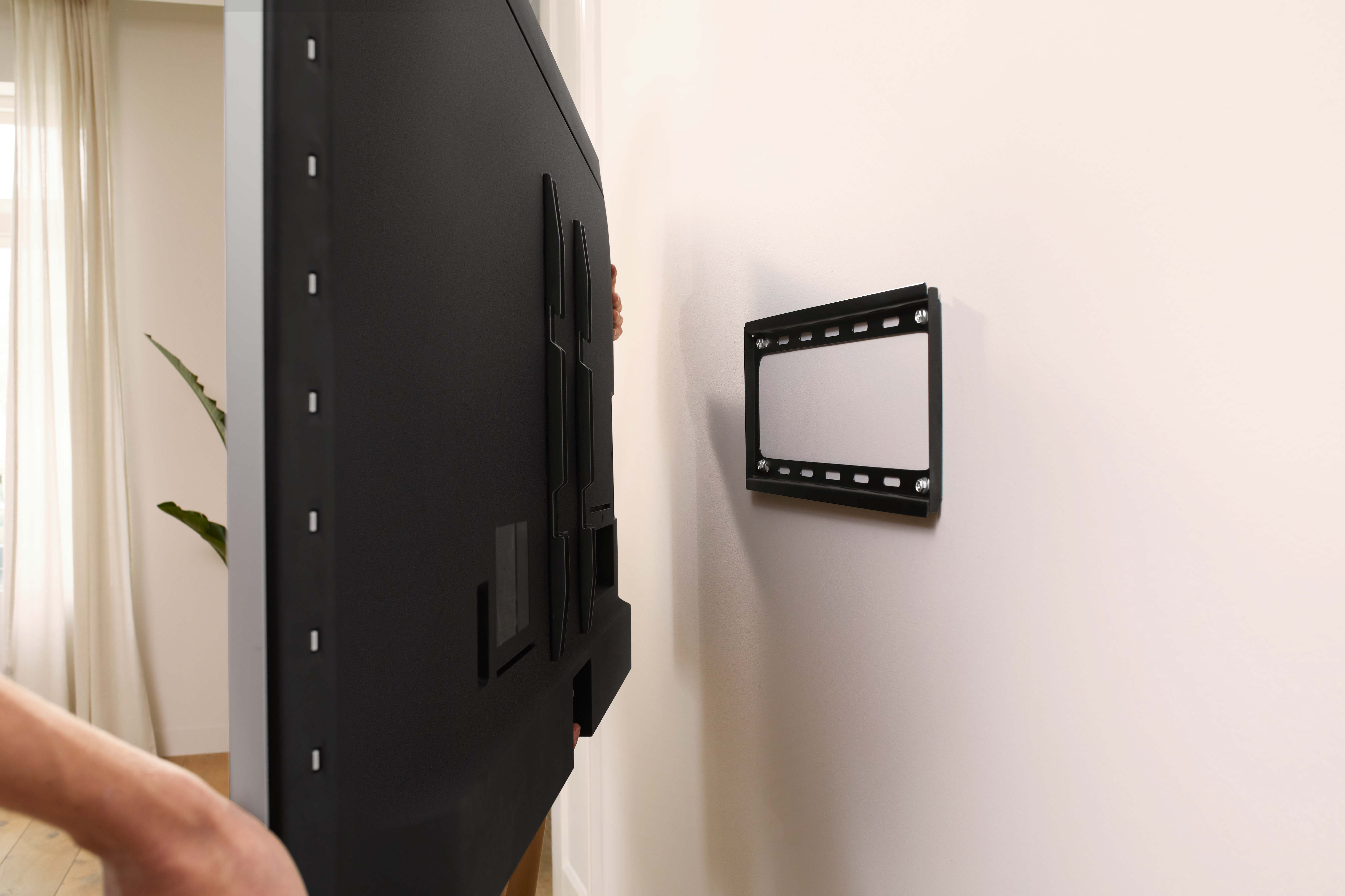 Een persoon houdt een zwarte tv vast terwijl deze aan een muur wordt gemonteerd, met de achterkant van de tv en de montagebeugel.