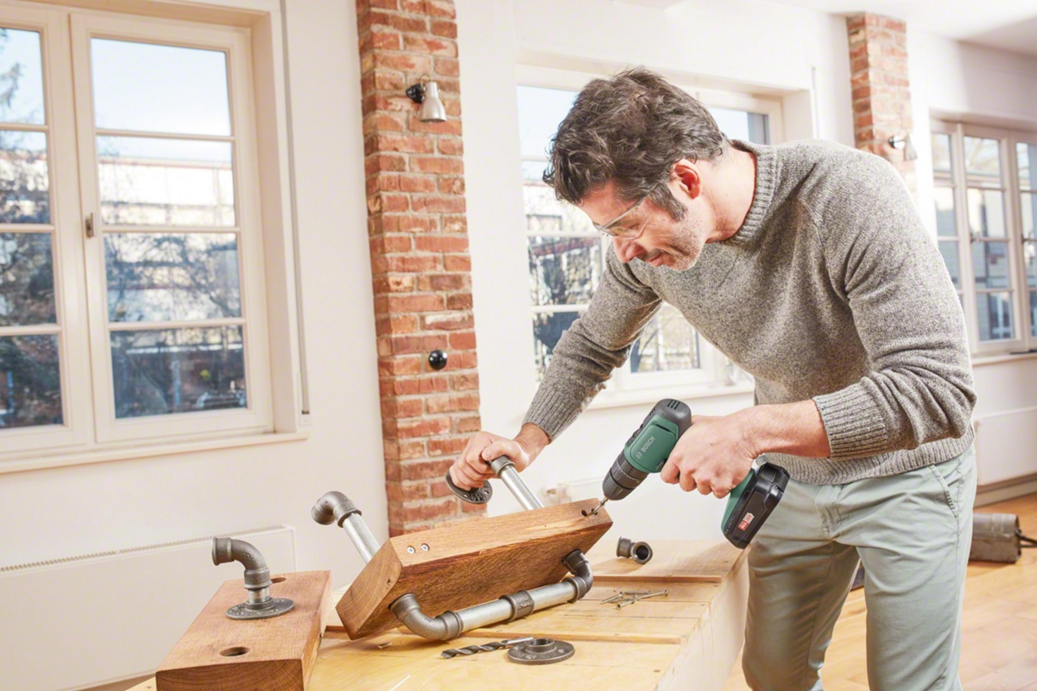 Hombre taladrando madera con tuberías, usando gafas de seguridad.