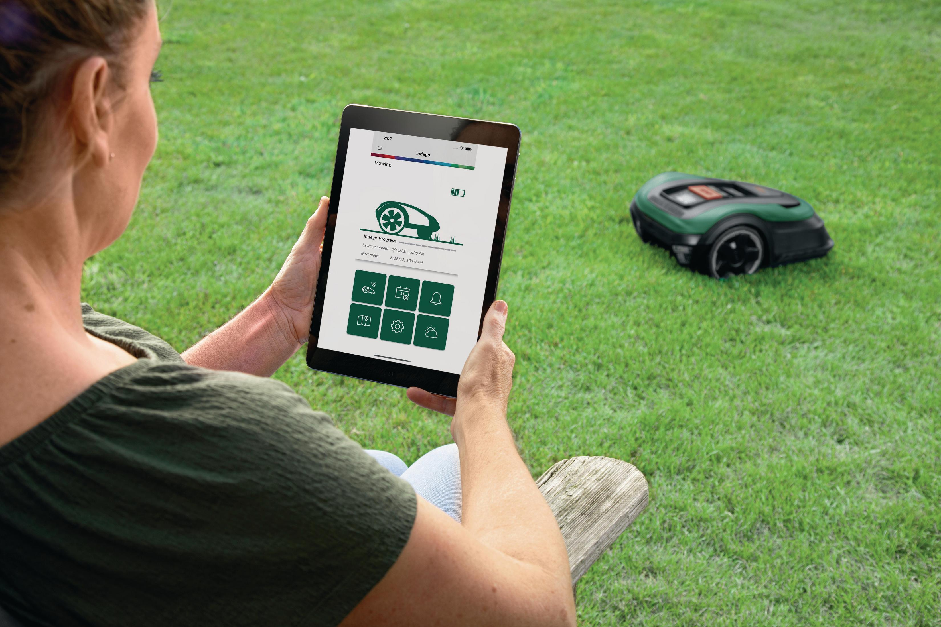 Kobieta używająca tabletu do sterowania kosiarką automatyczną. Kosiarka jest na zielonej trawie.