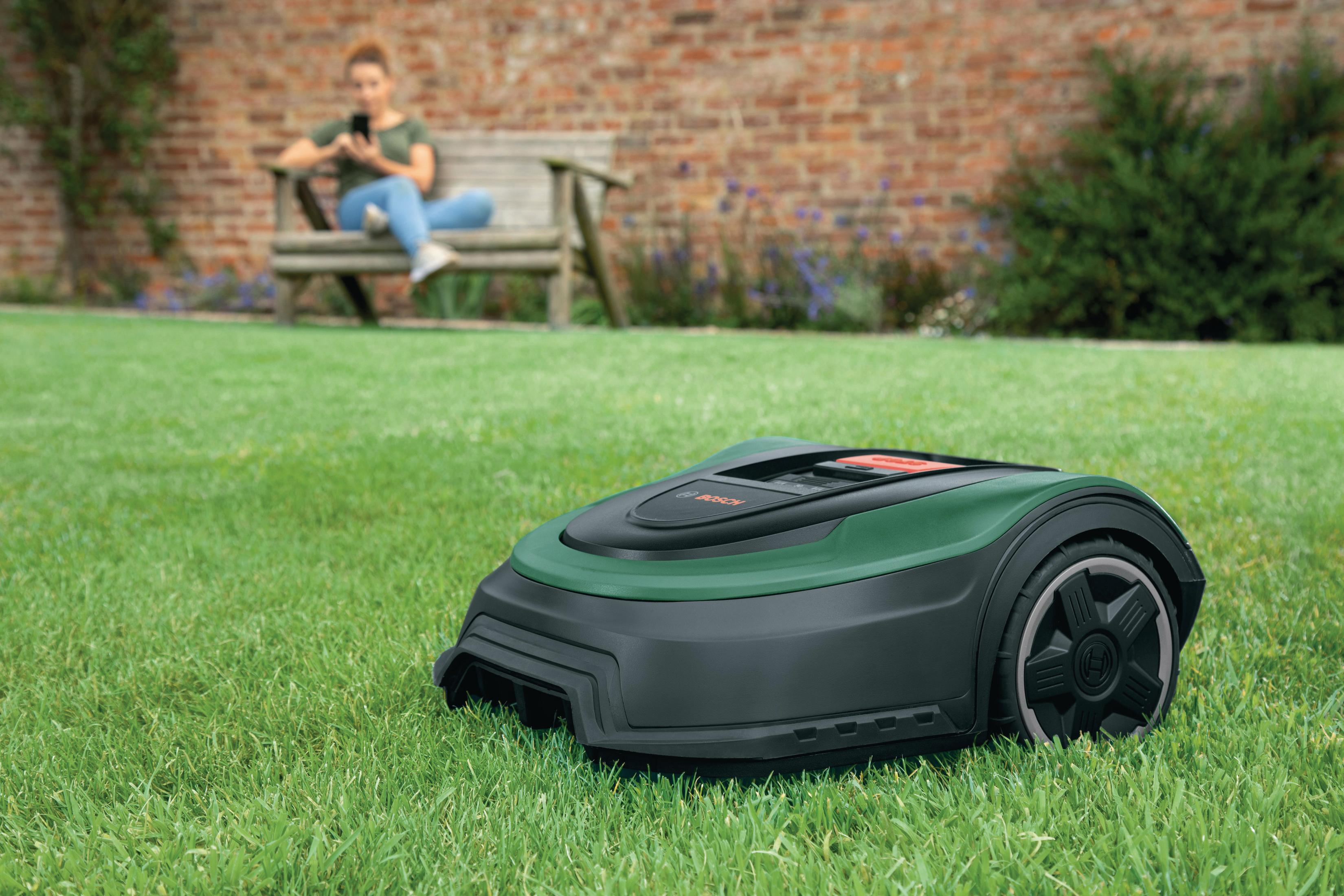 Zielona i czarna kosiarka robotyczna na trawie, kobieta na ławce w tle używa telefonu.