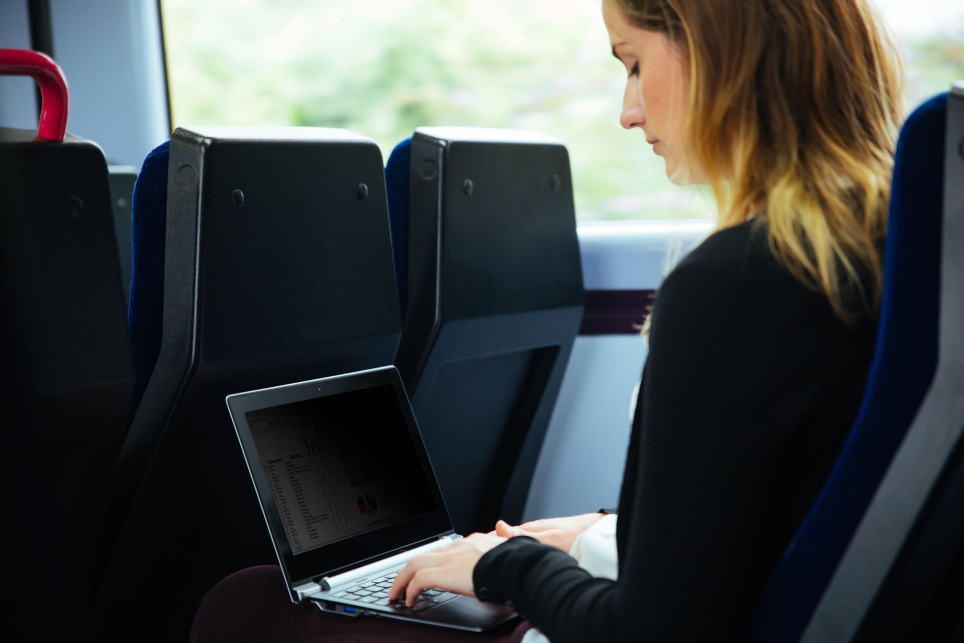 Eine Frau arbeitet in einem Zug an einem Laptop. Sie trägt eine schwarze Jacke und der Laptop-Bildschirm ist dunkel.