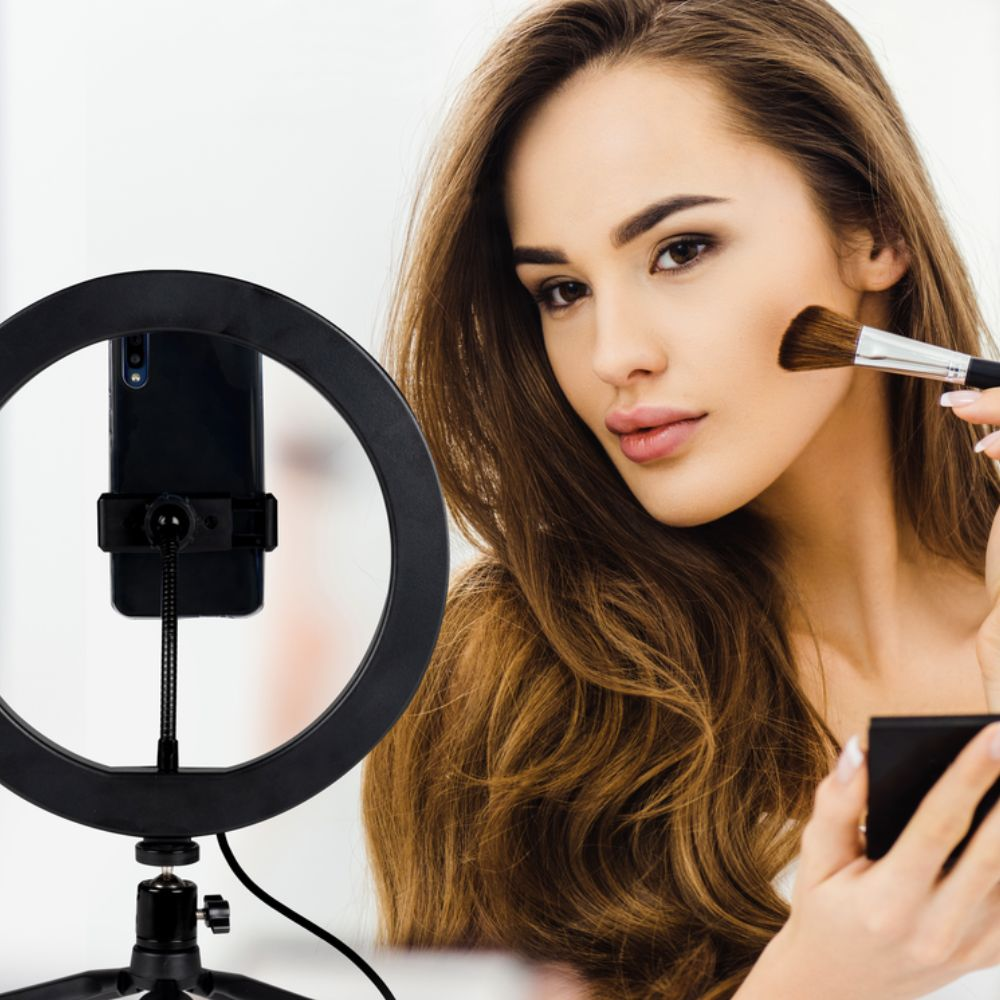 Une femme applique du maquillage avec un pinceau et un poudrier près d'un anneau lumineux et d'un téléphone.