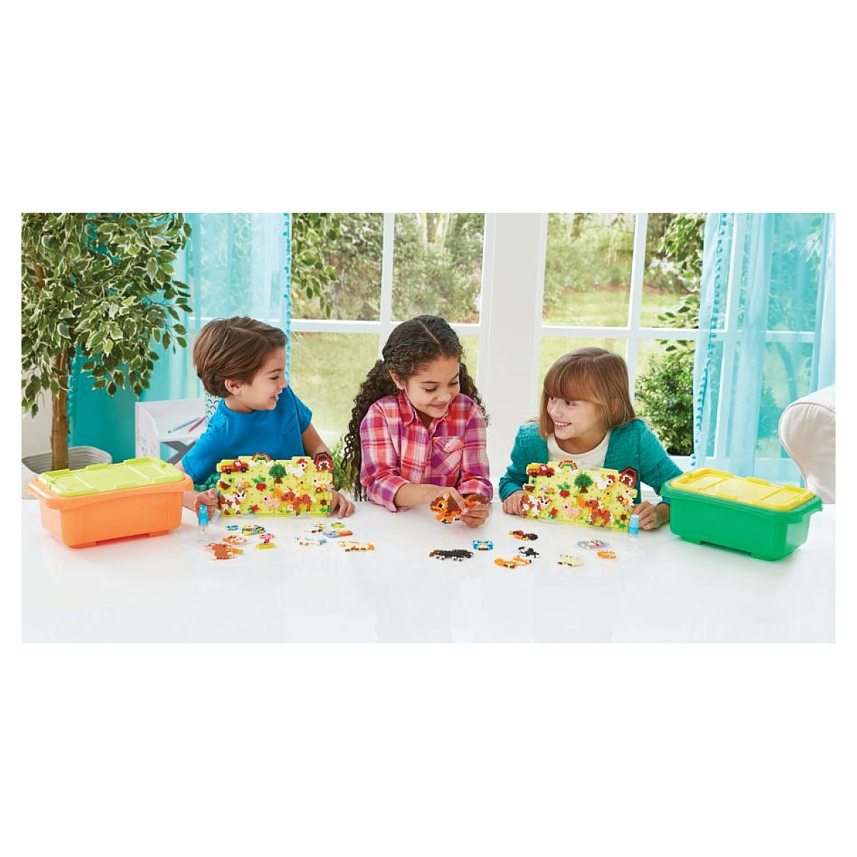 Drei Kinder an einem weißen Tisch spielen mit bunten Objekten. Auf dem Tisch befinden sich zwei orangefarbene und eine grüne Aufbewahrungsbox.