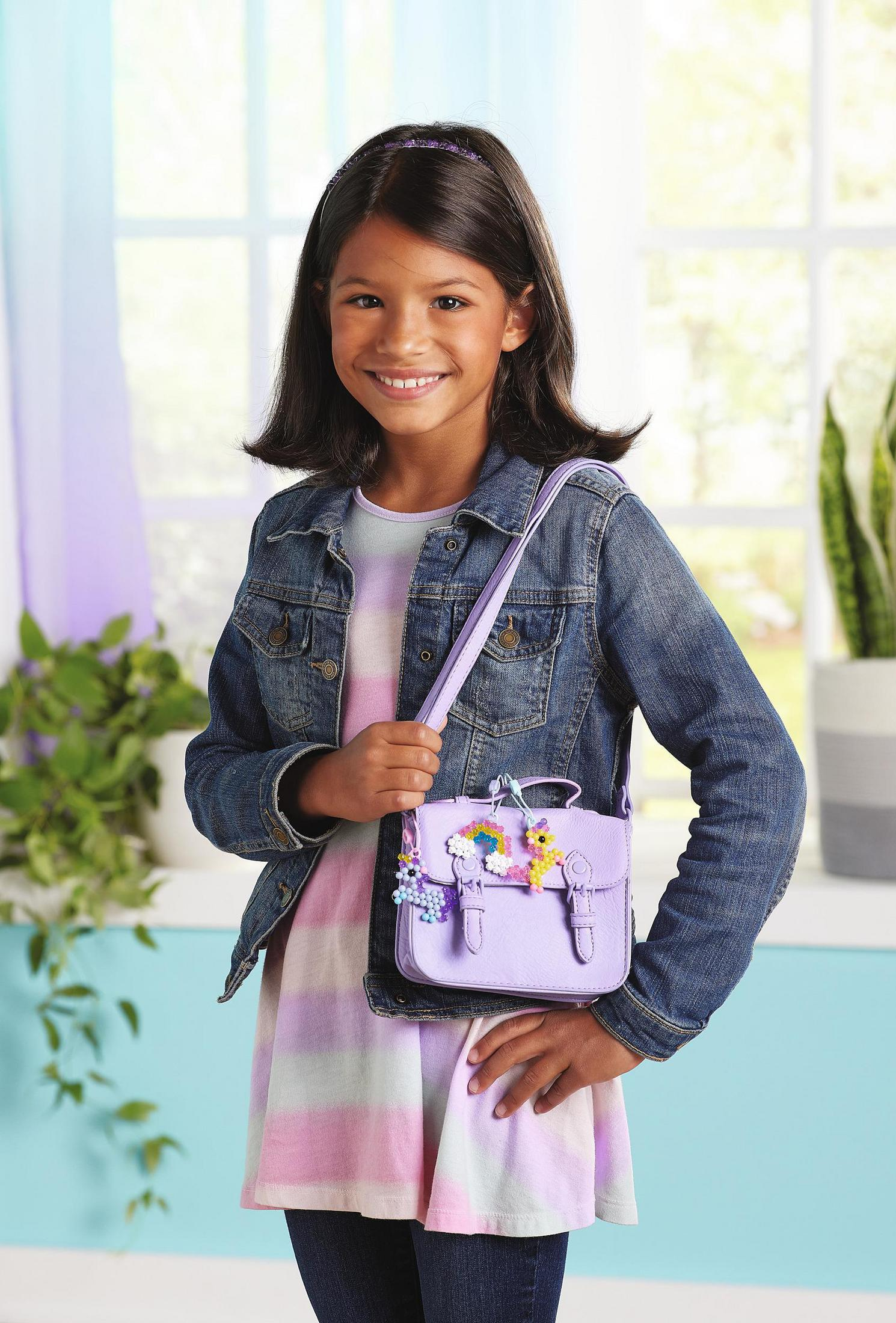 Ein junges Mädchen lächelt und hält eine lila Handtasche mit Blumenakzenten. Sie trägt eine Jeansjacke und ein gestreiftes Oberteil.