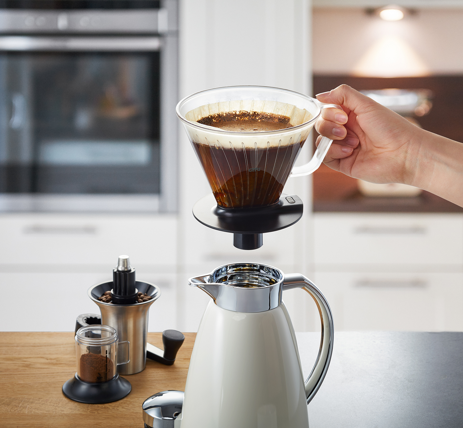 Hand gießt Kaffee in eine weiße Karaffe. Kaffeemühle und Bohnen im Hintergrund auf einem Holztisch.