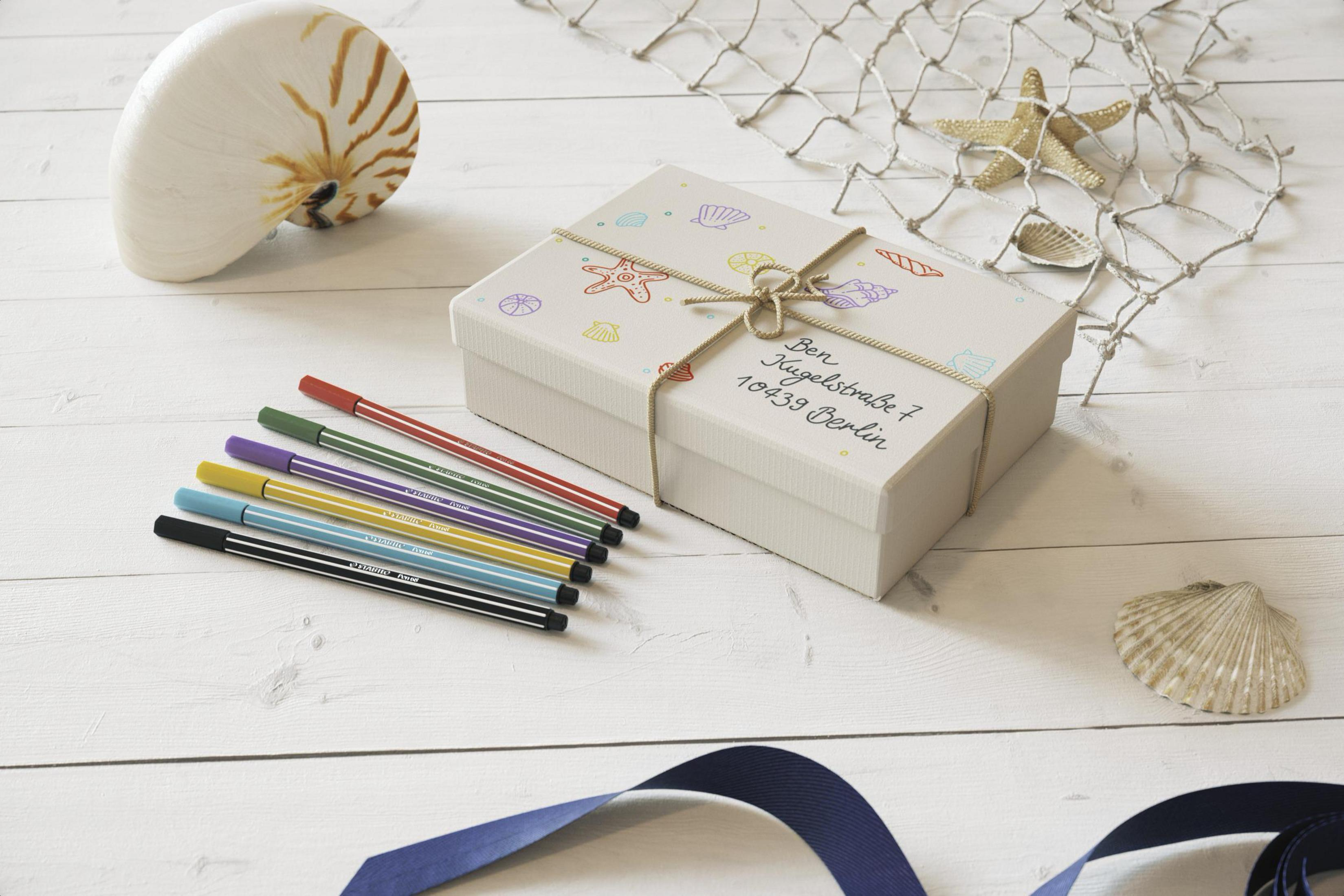 Geschenkbox mit Muscheln, Seesternen und bunten Stiften auf weißer Holzoberfläche, mit blauem Band.