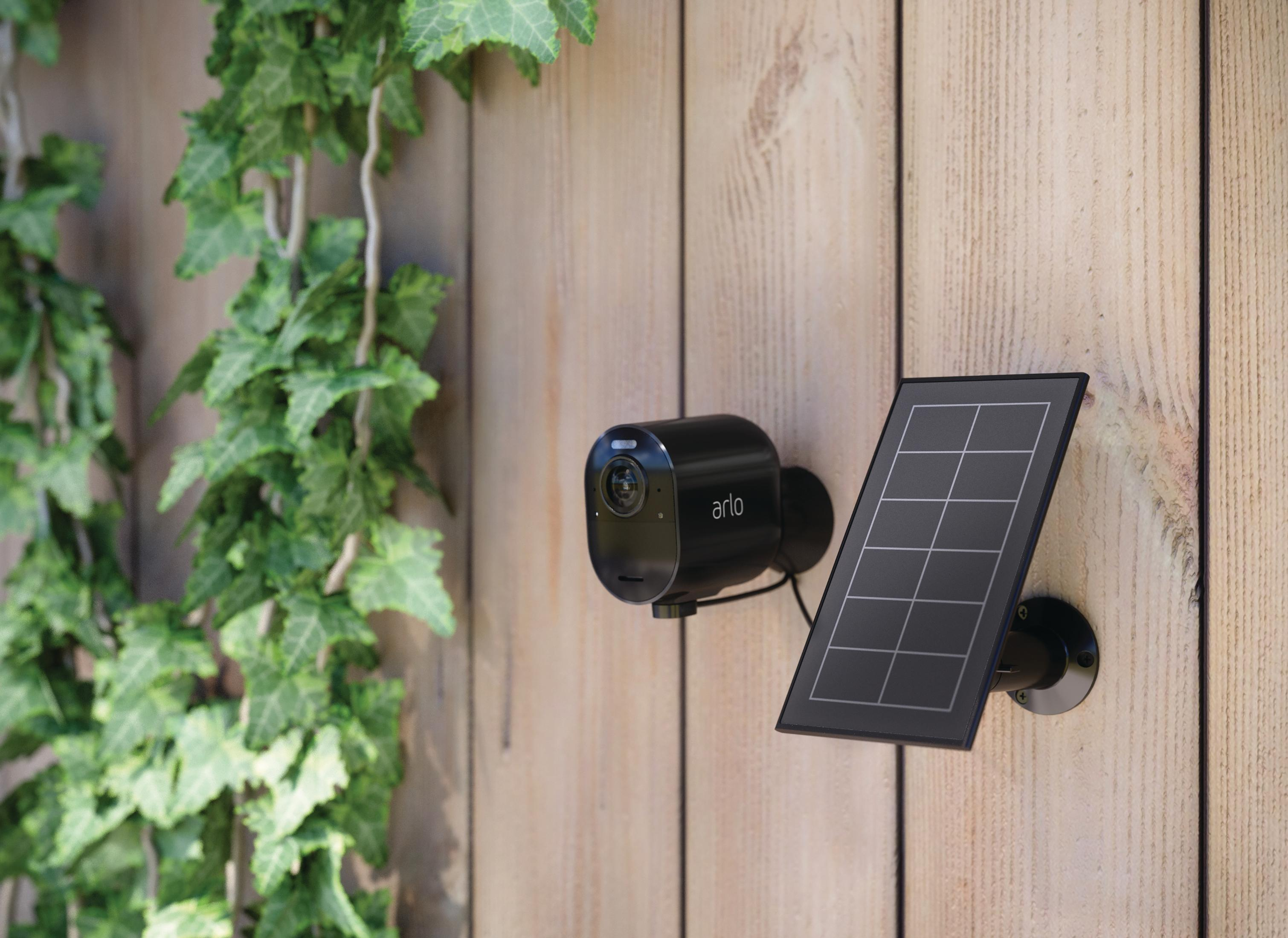 Schwarze Überwachungskamera und Solarpanel an einem Holzzaun mit Efeu befestigt.