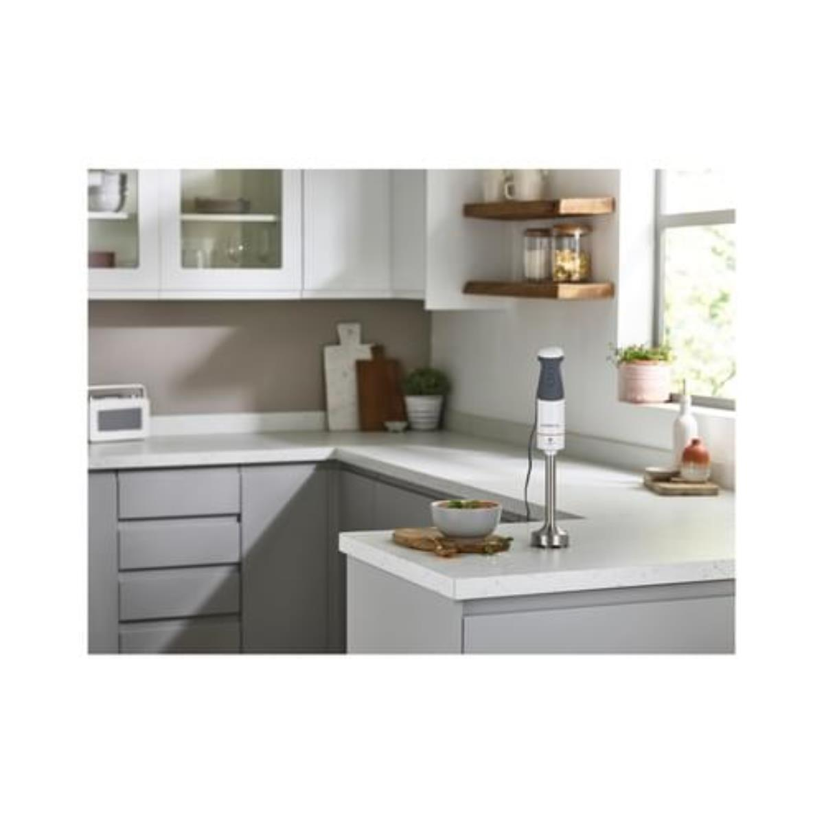 Cucina con un frullatore sul bancone. Armadi grigi, piani di lavoro bianchi. Mensole in legno.