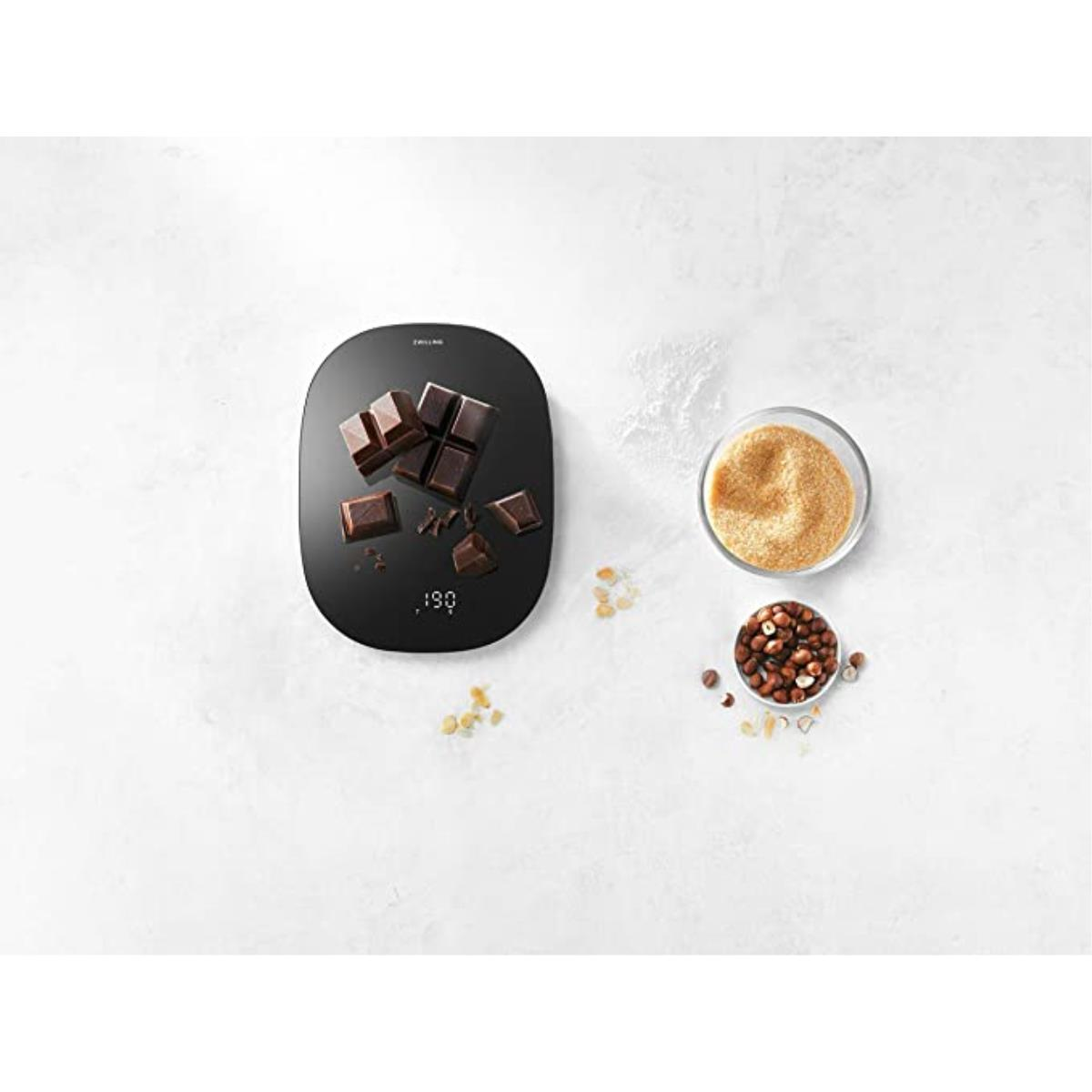 Czarna cyfrowa waga kuchenna z rozrzuconą czekoladą, cukrem i orzechami laskowymi.