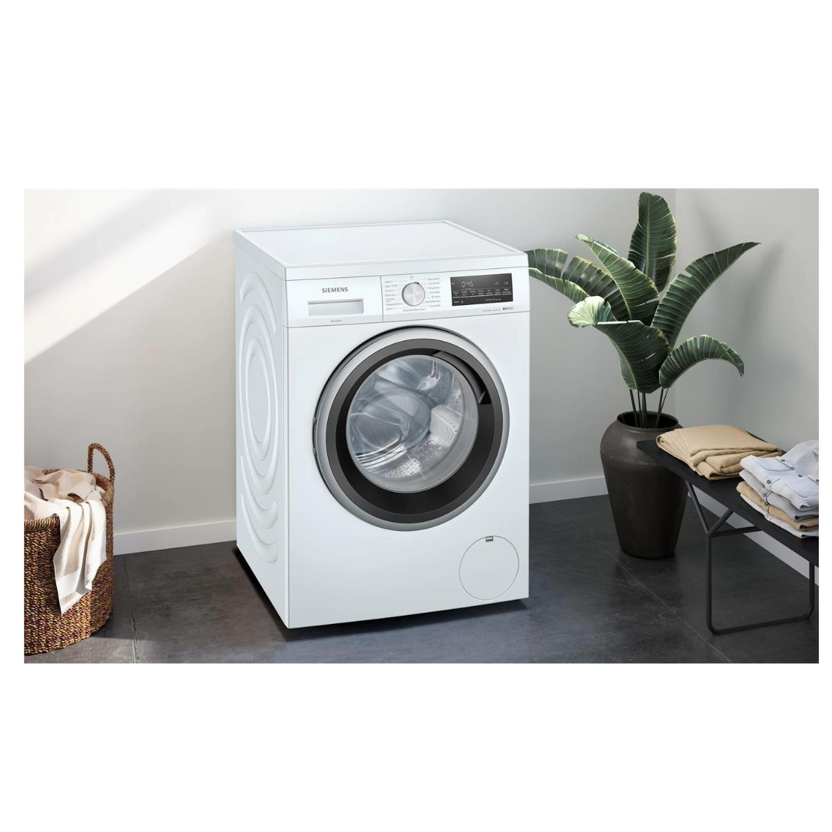 Biała pralka w pokoju z koszem na pranie i rośliną w doniczce.