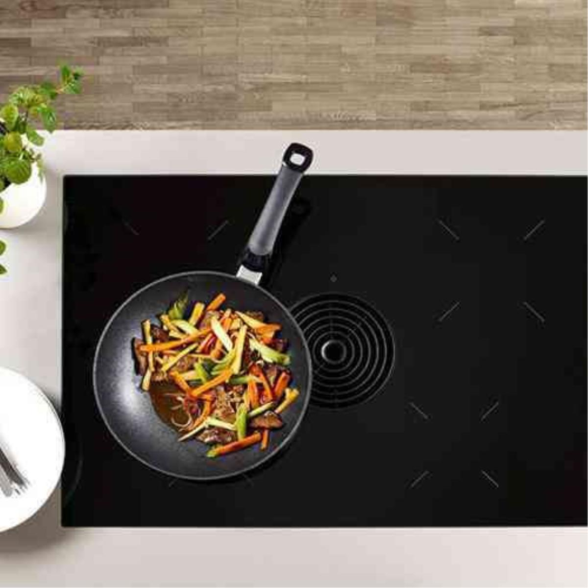 Una padella con verdure su un piano cottura nero con uno sfiato rotondo.