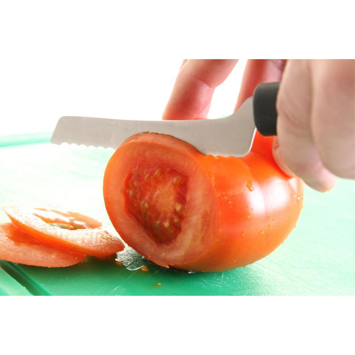Hand schneidet eine Tomate mit einem gezahnten Messer auf einem grünen Schneidebrett.