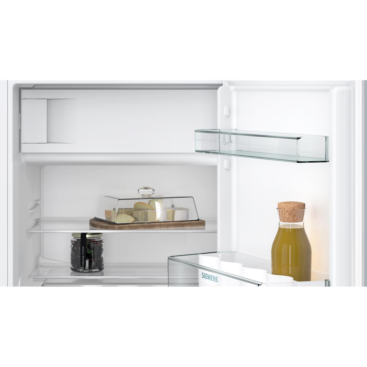 Binnenkant van een koelkast met items: kaas, een pot, een fles vloeistof en glazen planken. Wit interieur.