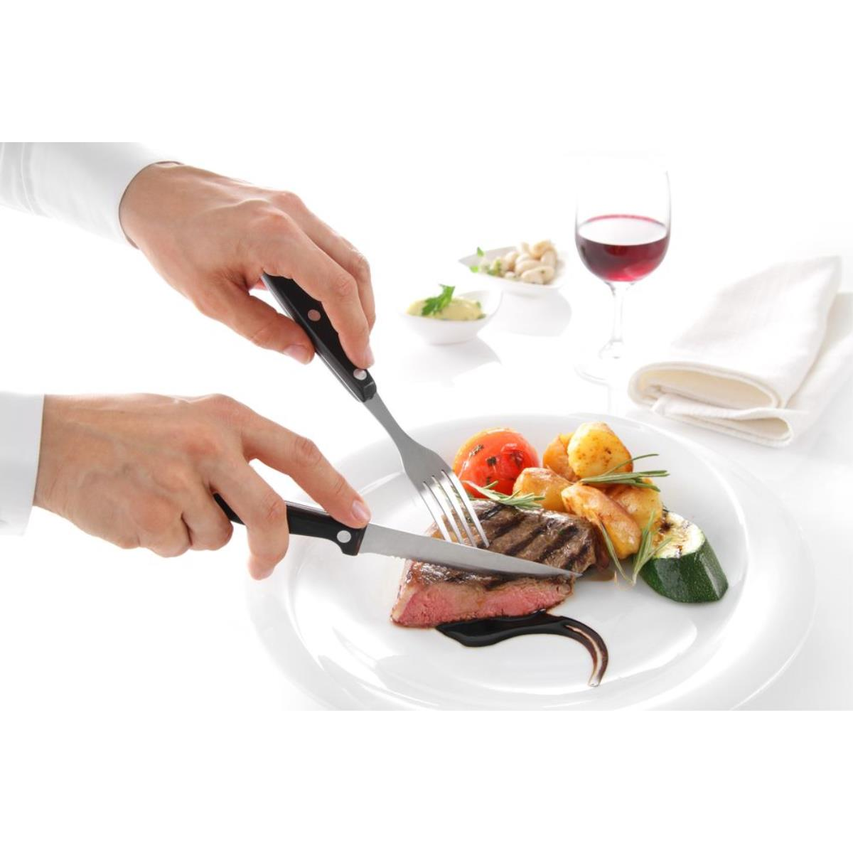 Hände schneiden Steak auf weißem Teller. Teller hat gegrilltes Gemüse und Weinglas.