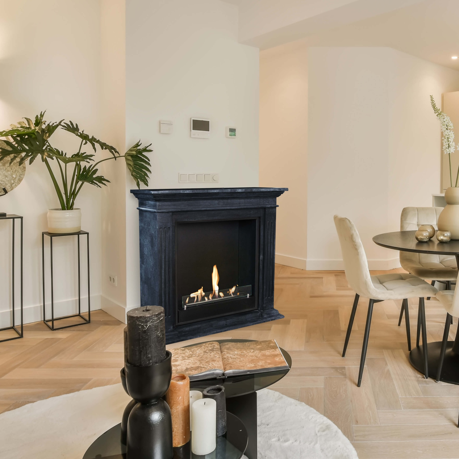 Een woonkamer met een open haard, een ronde tafel, stoelen en decoratieve items. Een plant staat bij de open haard.