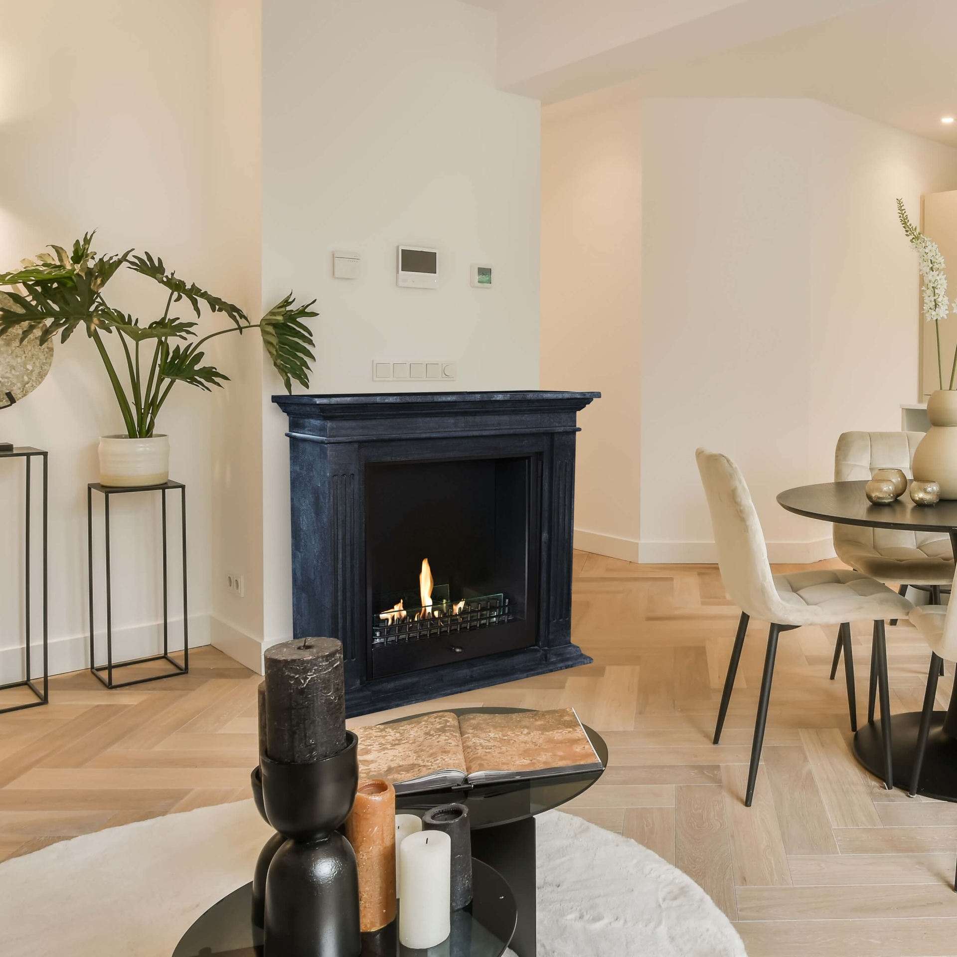 Een woonkamer met een open haard, een ronde tafel met stoelen, een plant en kaarsen op een salontafel. De vloer is parket.