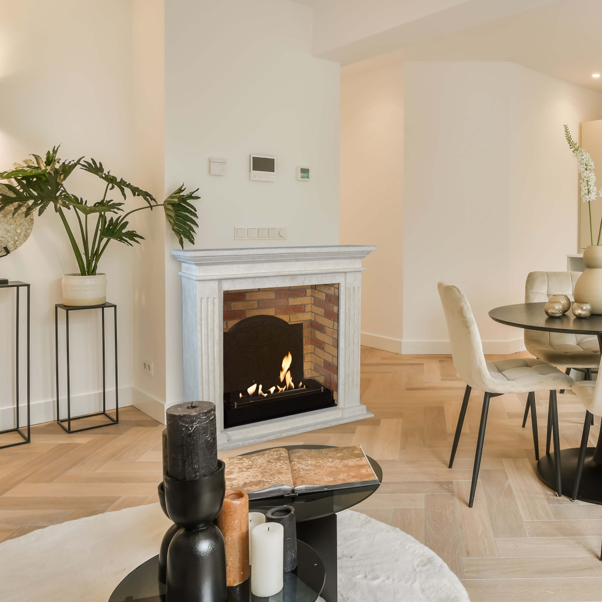 Een woonkamer met open haard, eettafel en decoratieve kaarsen. Een plant staat op een standaard. Houten vloeren.