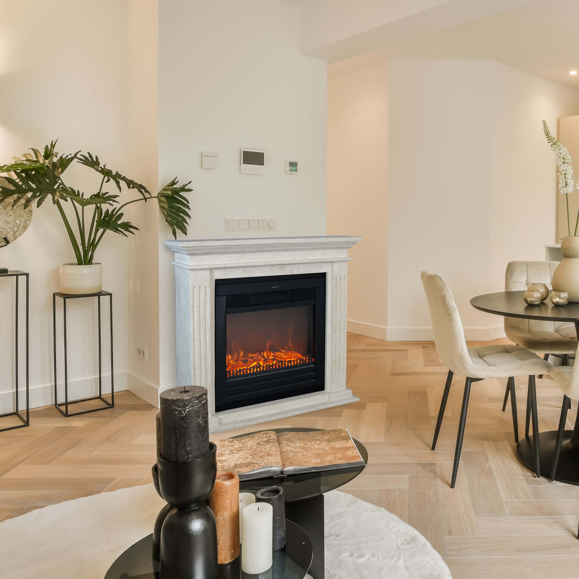 Een woonkamer met een open haard, kaarsen, ronde tafel, stoelen en planten. De vloer is van hout. De open haard heeft een vuureffect.